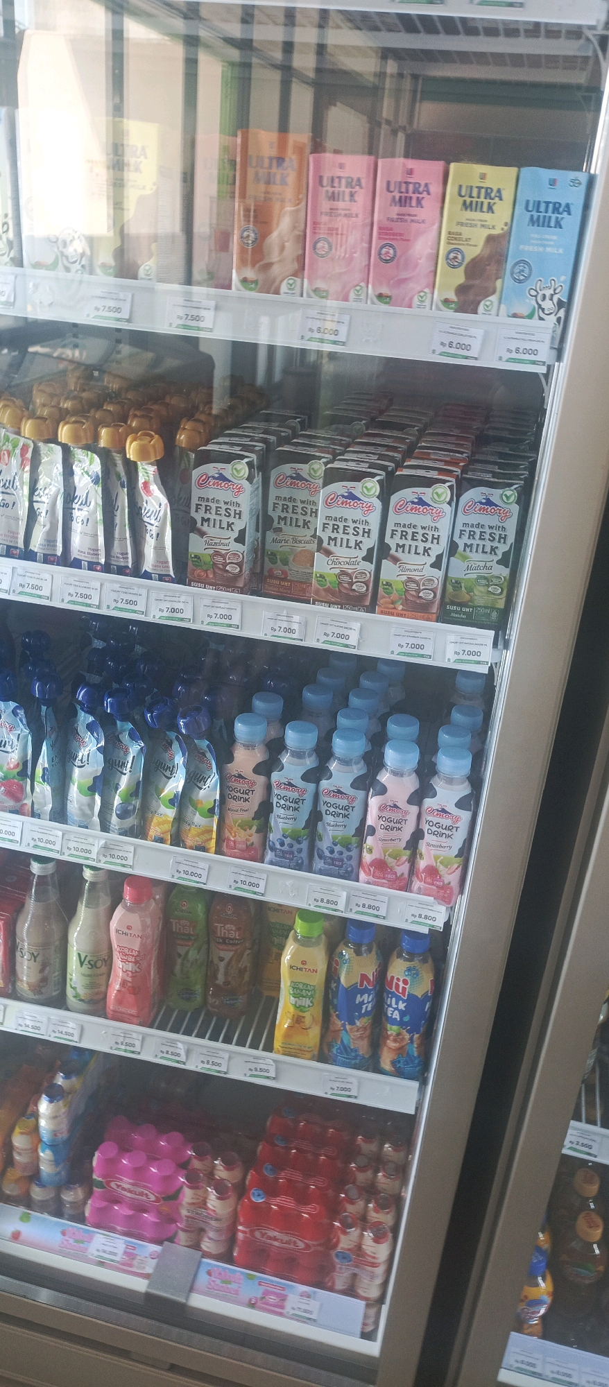 The image shows the interior of a refrigerated display case in what appears to be a convenience store or minimarket. The display is stocked with a variety of milk and yogurt products, predominantly packaged in cartons and bottles.
On the top shelf, several cartons of "ULTRA MILK" are visible in various flavors including orange, pink, brown, and blue packaging. Below this, a shelf displays pouches of yogurt with yellow caps, and further down, cartons of "Cimory made with FRESH MILK" are arranged in rows, featuring flavors like Hazelnut, Marie Biscuits, Chocolate, Almond, and Matcha.
The second shelf from the bottom contains bottled yogurt drinks from "Cimory" in flavors like Mixed Fruit, Blueberry, and Strawberry, along with other bottled beverages. The bottom shelf is filled with more bottled drinks and packs of Yakult.
The overall environment is clean and well-organized, typical of a retail setting. The presence of pricing labels indicates that these items are for sale. The lighting is bright, suggesting the interior of a store with overhead lights or internal illumination of the display case. There are no people visible in the image. The context implies this is a retail location in Lamongan, Indonesia, given the currency labels (Rp) and product branding.