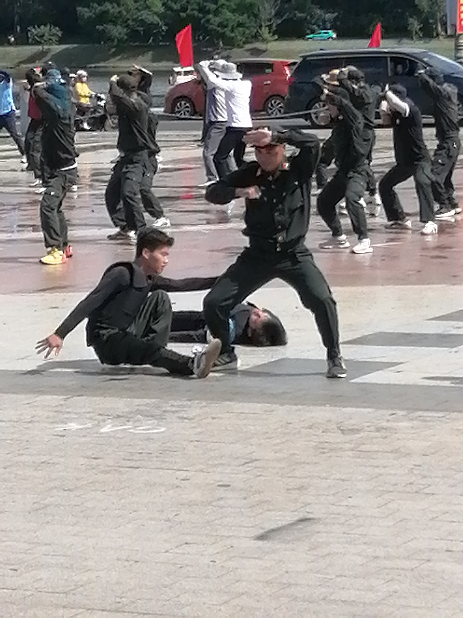 A group of people, dressed in black uniforms and vests, are performing a synchronized dance routine on a wet, paved outdoor area. The location is identified as Da Lat City, Vietnam, a detail that might suggest the performance is part of a cultural event or demonstration. Several individuals are captured in dynamic poses, with some in mid-air leaps and others in low stances. One person is lying on the ground, seemingly part of the choreography. In the background, a red car and a black SUV are visible, along with trees and a body of water, indicating an urban or park-like setting. The ground is reflective, suggesting recent rain or a water feature. The overall scene depicts a lively and energetic performance.