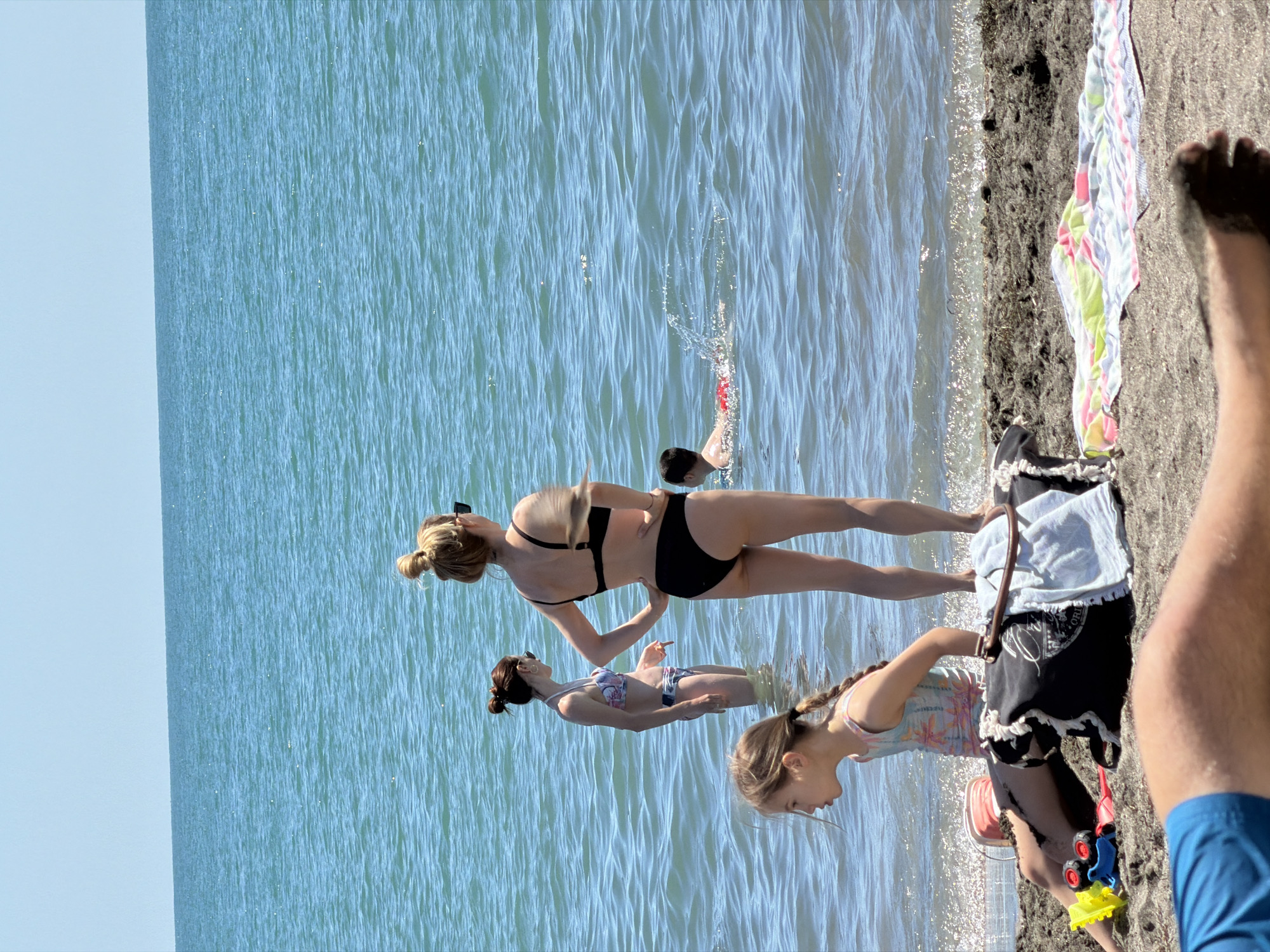 This media file captures a sunny day at the beach in Burgas, Bulgaria, with the sea on the left and the sandy shore on the right. Three women are in the water, with one woman in the foreground wearing a black bikini and sunglasses. Behind her, another woman in a patterned bikini is partially submerged in the water. A young girl, also in the water, is visible in the lower right of the frame, wearing a patterned swimsuit. On the shore, a colorful beach towel is laid out, and a child's red and blue toy truck is visible. The time of day appears to be midday, given the bright sunlight casting shadows and highlighting the water's surface. The overall mood is relaxed and leisurely, consistent with a typical beach outing. The clear blue sky and calm water suggest pleasant weather conditions.