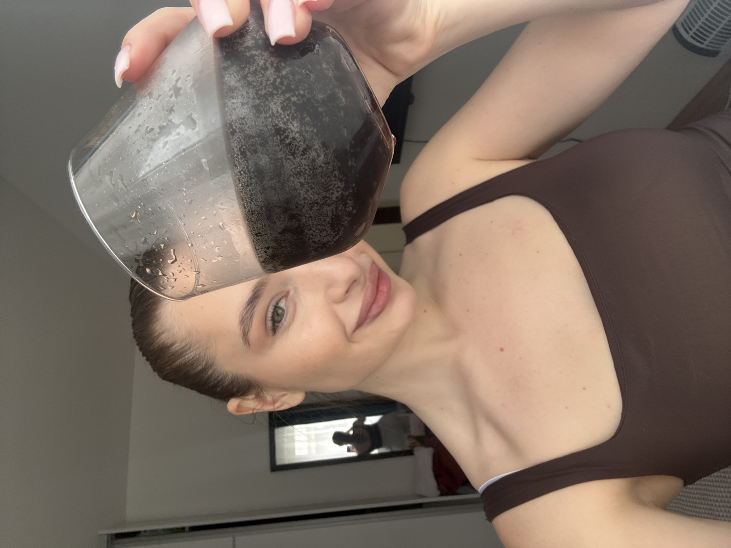 This portrait-oriented image, rotated 90 degrees counter-clockwise, captures a young woman holding a glass of a dark, carbonated beverage. The woman, likely in her 20s, has light skin, green eyes, and her brown hair is pulled back. She sports light pink manicured fingernails. She is smiling softly and looking towards the viewer, appearing relaxed and content.
She is wearing a dark brown, form-fitting top or bodysuit with a distinctive cut-out detail along the shoulder and side. Her arm is raised, holding a clear glass cup that is visibly cold with condensation and water droplets on its exterior. The glass is filled with a dark, effervescent liquid, indicating a carbonated drink. The cup is tilted, suggesting she is in the process of drinking or has just taken a sip.
The background shows an indoor setting, presumably a room with light-colored walls. A dark-framed object, possibly a mirror or picture frame, is visible behind her, reflecting a faint figure. A white fan or vent is also partially visible in the upper right portion of the image (when upright). The time of day appears to be daytime due to the ambient light. While no specific visual cues within the image confirm the location, it is contextually set in Budva, Montenegro. There is no visible text.