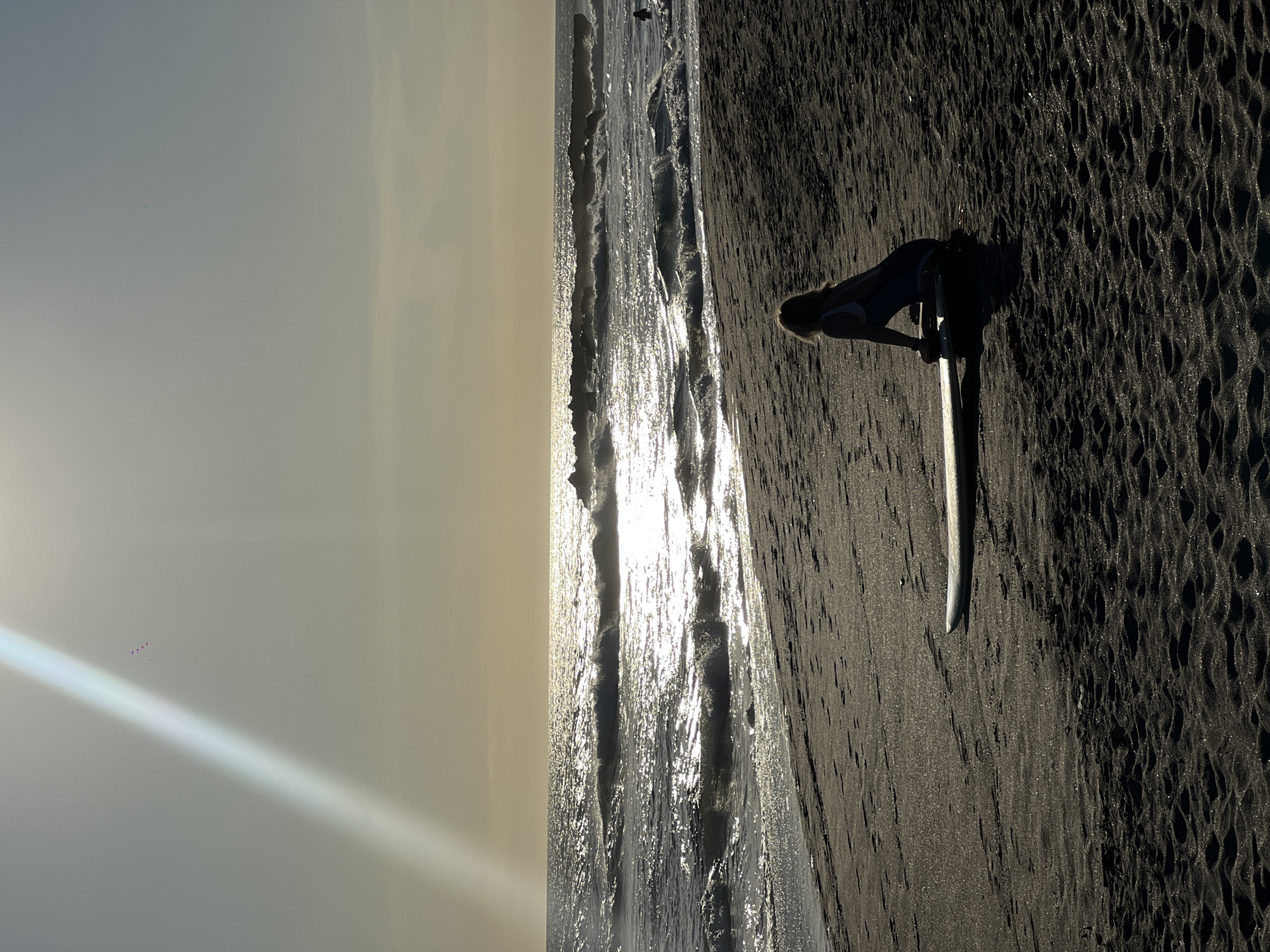 Here's a description of the image:
**Location:** Appears to be a beach in North Kuta, Indonesia, based on the dark sand characteristic of that region.
**Scene:** A woman is standing on the beach, her head bowed toward a surfboard. The surfboard is upright, seemingly positioned in the sand. Waves are visible in the background, reflecting the sunlight. The sand is dark. The lighting suggests a time around sunset or sunrise.
**People:** One woman is visible.
**Objects:** Surfboard, waves, sand.
**Actions/Interactions:** The woman is interacting with the surfboard. The waves are moving toward the beach.