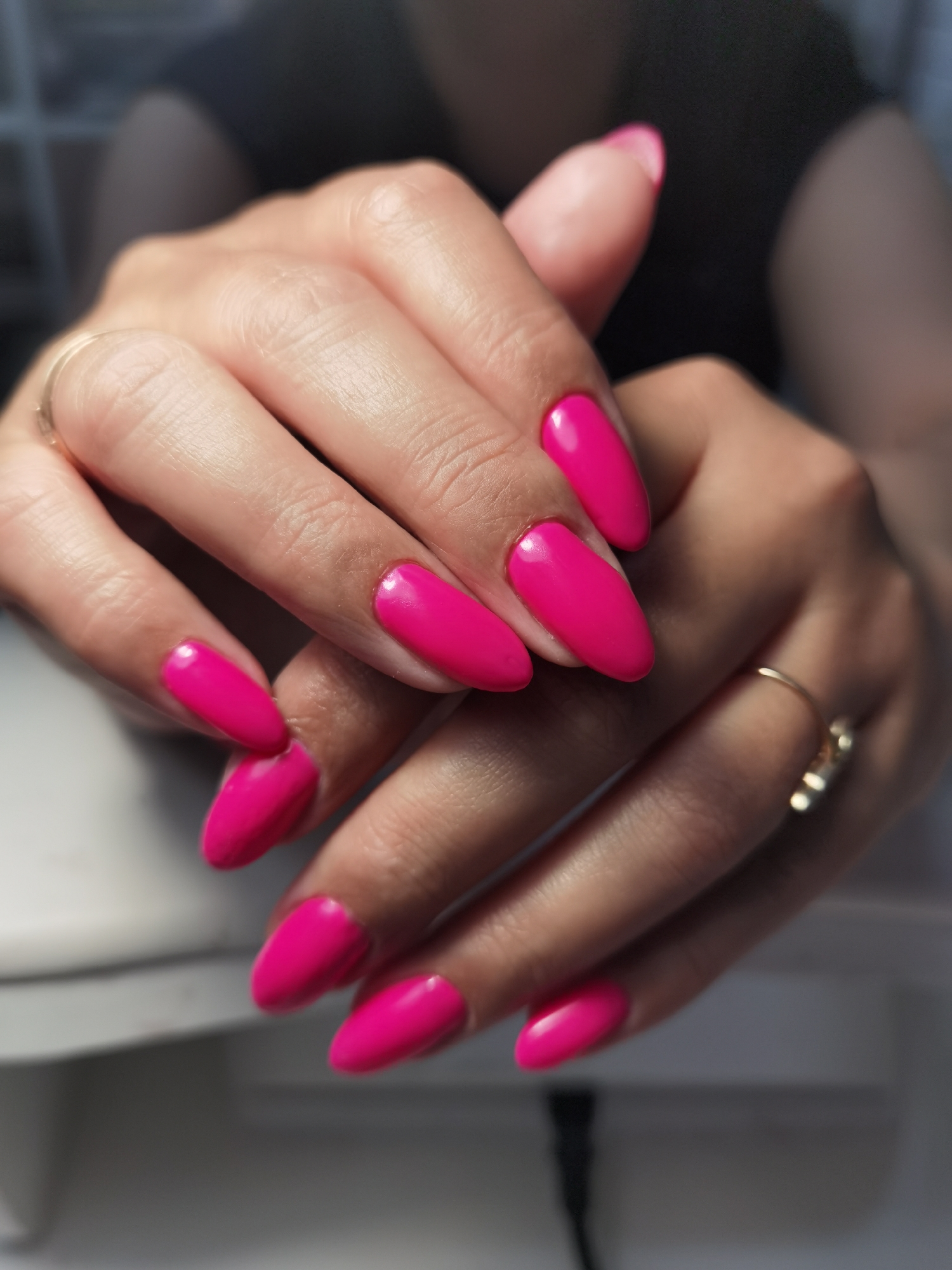 The image displays a close-up of a person's hands with bright, hot pink, oval-shaped nails, indicating a recent manicure. Both hands are clasped or resting one over the other, showcasing the vibrant nail polish and clean cuticles.
On the left hand, a simple, thin gold-colored band ring is visible on the ring finger. On the right hand, a more intricate gold-colored ring with small, possibly diamond-like, embellishments is worn on the ring finger. The skin tone of the hands is light to medium.
The person is wearing dark, out-of-focus clothing, possibly a black t-shirt, visible in the background and partially behind the hands. The immediate foreground shows a blurry, light-colored surface, possibly a table or a nail drying lamp, and a dark cable or cord is faintly visible towards the bottom. The overall background is dark and heavily blurred, suggesting an indoor setting, likely a nail salon or a well-lit home environment in Chaikovsky, Russia, where the manicure was performed. There is no discernible text in the image.