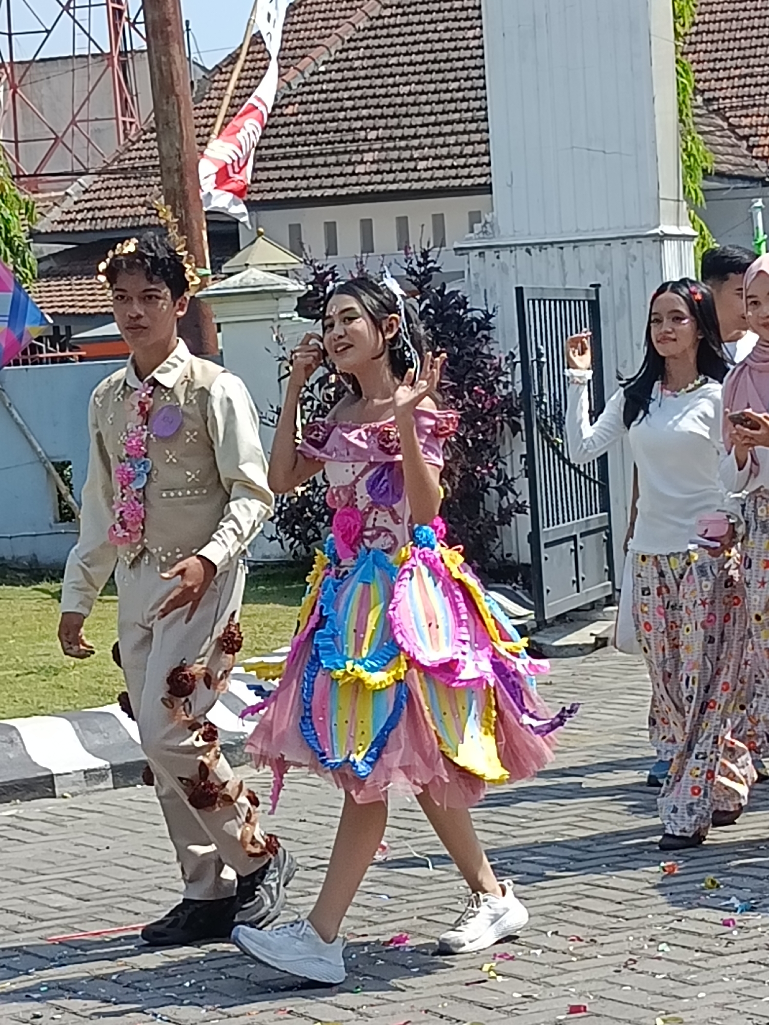 In Kediri City, Indonesia, a festive parade is underway as two individuals, a young man and a young woman, walk side-by-side on a paved street. The young man, wearing a light-colored vest over a collared shirt and matching pants, has a decorative floral garland around his neck and is adorned with small brown floral accents on his pants. A golden laurel wreath rests on his head. To his right, the young woman is dressed in a vibrant, multi-colored, ruffled dress. Her dark hair is adorned with white ribbons.
Behind them, other participants are visible. A young woman in a white long-sleeved shirt and patterned pants walks, holding a small pink purse. Further behind, another woman in a light pink hijab and patterned pants is also present. The street is decorated with scattered confetti, suggesting a celebration. Buildings with tiled roofs form the backdrop, and a tall wooden pole with a red and white flag attached stands to the left of the young man. The scene appears to be taking place in an outdoor urban environment.