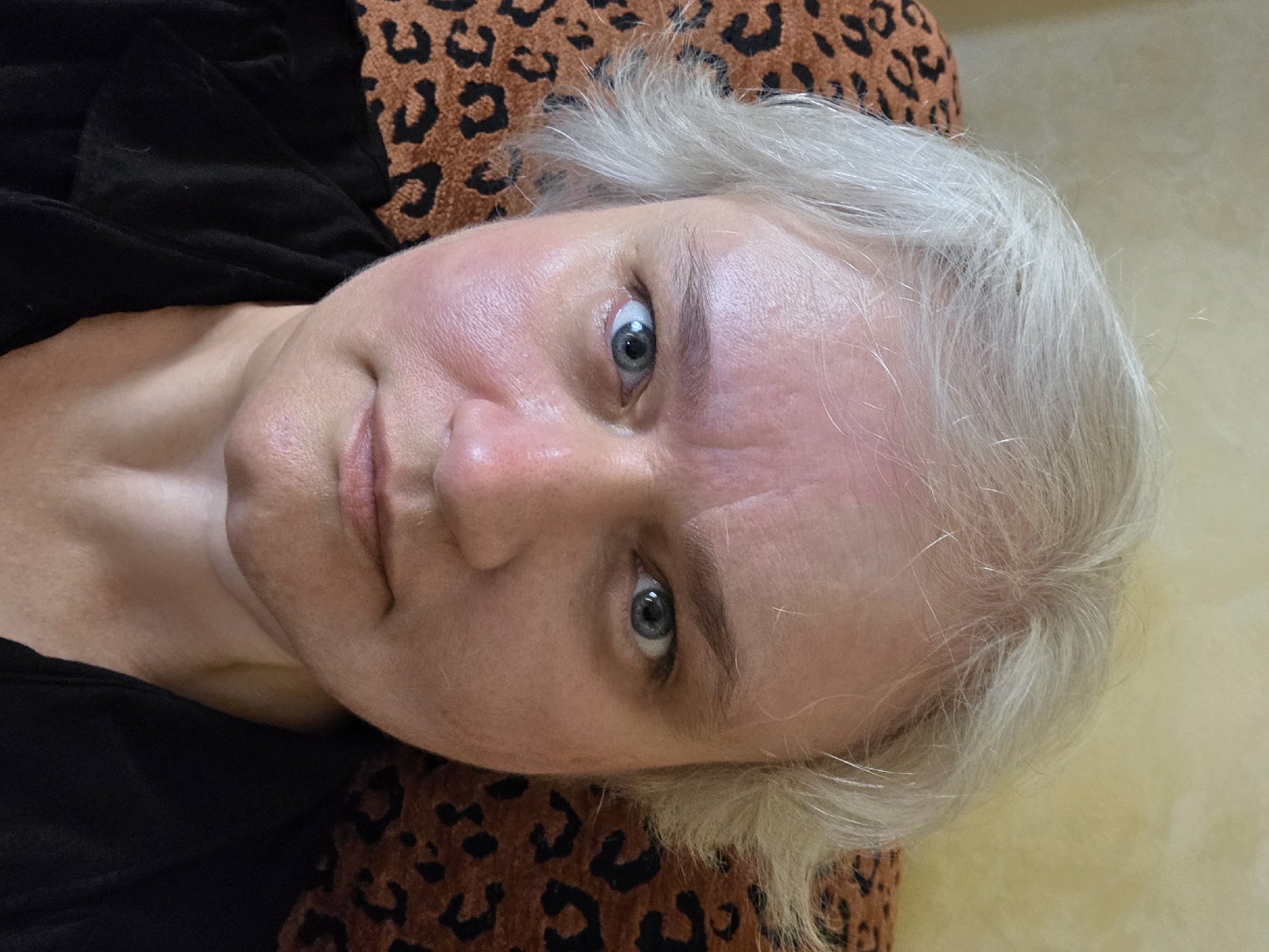 A medium close-up shot features a mature woman with light skin, gray hair, and blue eyes. The subject is positioned against a background featuring a leopard print design and a plain light-colored surface. She wears a dark-colored top. The image composition presents a portrait-style view of the woman's face and upper body. There is no contextual information available that would place this scene in Hurghada, Egypt.