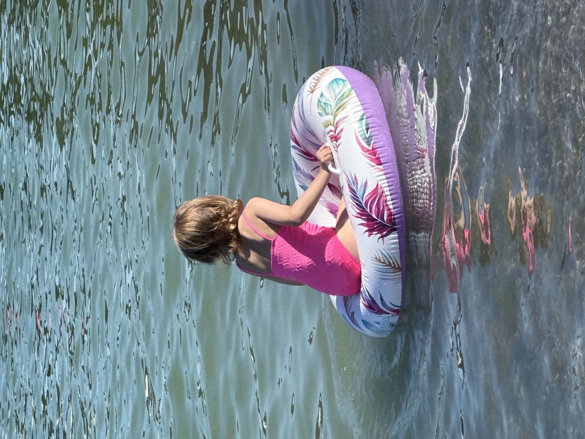A young girl in a pink swimsuit is playing in the water with a colorful inflatable ring. The ring is decorated with a tropical leaf pattern in shades of purple, green, and pink. The girl is holding onto the ring with both hands, and her body is partially submerged in the water. The water is clear and reflects the sunlight, creating a shimmering effect. The scene appears to be a sunny day at a lake or swimming pool. The girl's pose suggests she is enjoying herself. The image is taken from a high angle, looking down at the girl and the water. There is no visible text in the image.