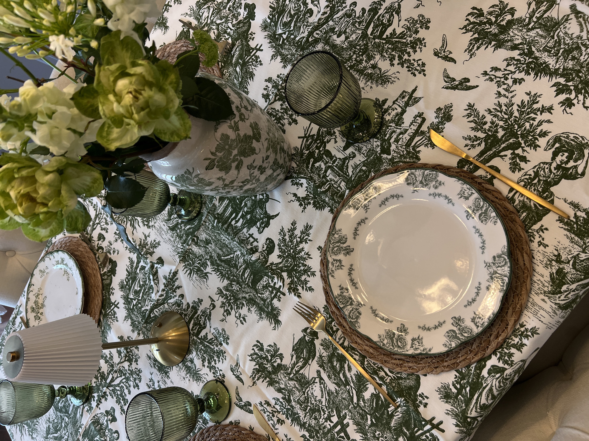 An elegant dining table is set indoors, viewed from an overhead perspective. The table is draped with a white tablecloth featuring a detailed green toile de Jouy pattern, depicting pastoral scenes with figures, trees, birds, and various landscape elements.
At the center, a white ceramic vase with a matching green floral design holds a fresh bouquet of white and green flowers, including prominent ruffled green hellebores and other delicate white blossoms. Several place settings are arranged around the table. Each setting consists of a round, woven straw or wicker placemat topped with a white dinner plate. These plates have a scalloped edge and a beautiful green floral pattern along their rims.
Beside the plates, gleaming gold-colored cutlery, including forks and knives, is neatly laid out. Green-tinted, ribbed glass goblets or water glasses complement the green and white color scheme of the table. A small decorative lamp with a pleated white shade and a brass base is visible on the left side of the table, contributing to the sophisticated ambiance.
The table is meticulously set, suggesting preparation for a formal meal, brunch, or special gathering. No people are visible in the image, but the arrangement indicates an imminent event. The lighting appears to be soft and ambient, typical of an indoor setting. There are no specific visual cues within the image to indicate its location in Moscow, Russia, other than the provided context. No text is visible on any of the objects. The scene conveys a sense of elegance, refinement, and a welcoming atmosphere.