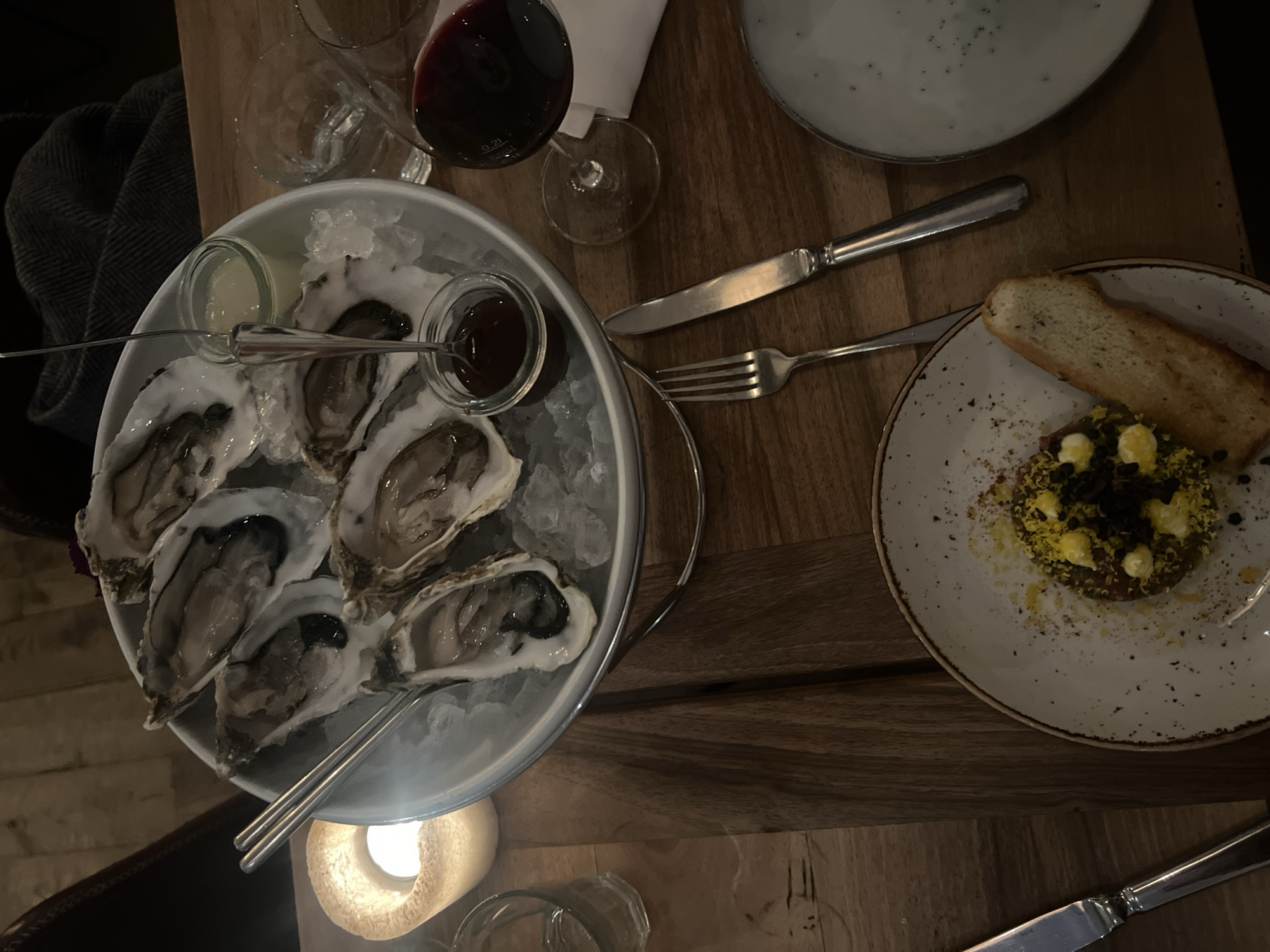 A dimly lit, top-down view of a wooden table arrangement. A plate of oysters on ice with two small sauce containers sits to the left. A glass of red wine, silverware, and two other plates are arranged around the oysters. One plate holds a dish with a slice of bread. A partially visible candle is in the bottom left corner. No people are visible. The location is likely a restaurant in Berlin, Germany, based on the dining setting.