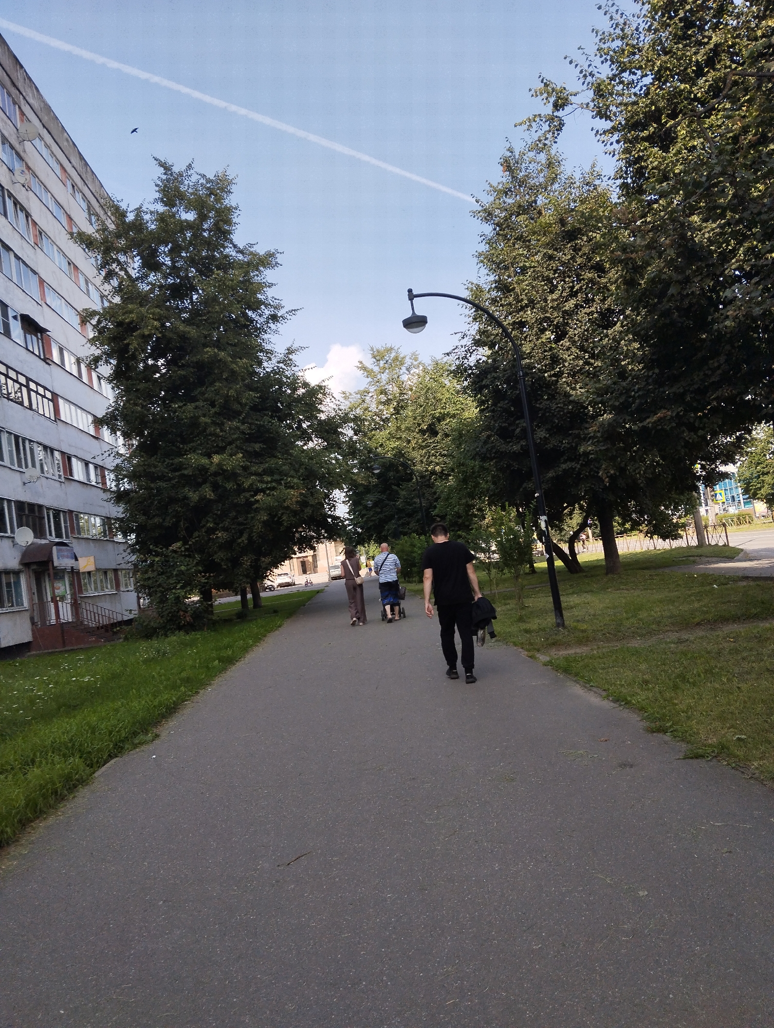 An exterior, eye-level, full shot shows a paved walkway lined with grass and trees, situated in front of a multi-story apartment building. A group of four people are walking on the path, with one person in the foreground walking away from the camera and holding a jacket. The apartment building is light grey with numerous windows. Trees with dense green foliage are present on both sides of the walkway. The sky is blue with a faint contrail from an airplane. A lamppost is visible on the right side of the walkway. The scene appears to be in a residential area in Kingisepp, Russia.