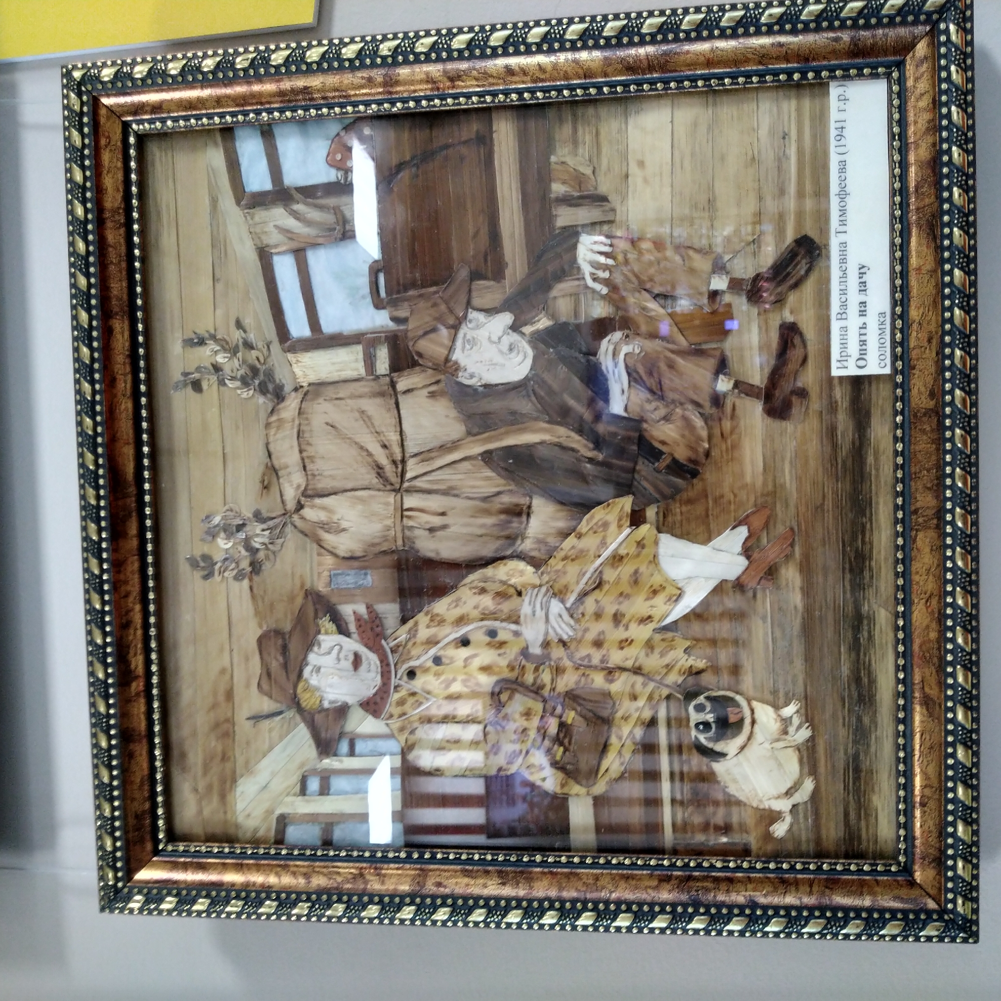 This is a framed artwork, likely a relief or collage, depicting two individuals and a dog in what appears to be a rustic interior setting, possibly a dacha, or country house. The artwork is made from straw (соломка).
In the foreground, a light-colored pug dog with a black nose and tongue is positioned to the left. Behind the dog, a woman is seated. She wears a brown hat, a patterned yellow coat with black spots, and white stockings. She holds a small brown handbag. To the right, a man is crouched. He wears a brown cap, a dark jacket, and pants. His hands are resting on his knees.
In the background, there are wooden walls and windows. A large, bundled object, possibly luggage, is situated between the two figures. Decorative sprigs of dried leaves are placed above this bundle.
A white label at the bottom right of the artwork reads: "Ирина Васильевна Тимофеева (1941 г.р.) Опять на дачу соломка". This translates to "Irina Vasilievna Timofeeva (born 1941) Back to the dacha straw."
The artwork is housed in an ornate frame with gold and black detailing. The overall scene evokes a sense of travel or a summer outing. The detail about the location in Krasnoye Selo, Russia is not directly evident from the image itself, but is provided as context.