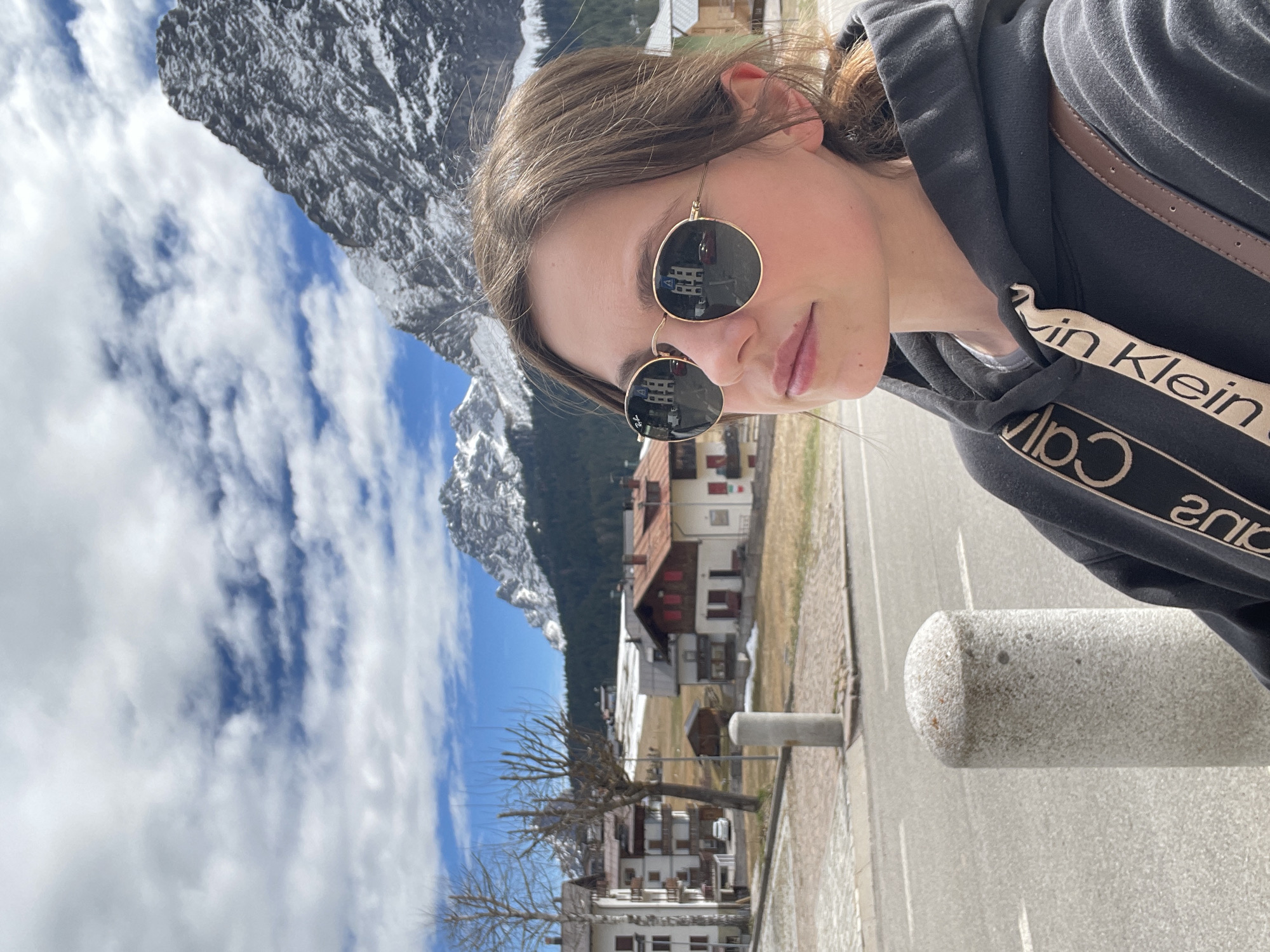 A photograph captures a person wearing a black Calvin Klein hoodie and sunglasses, with a mountainous backdrop and houses characteristic of Sappada, Italy. The mountains are snow-capped. The sky is blue with puffy clouds. Houses are visible in the reflection of the sunglasses. A cylindrical concrete bollard is partially visible in the foreground.