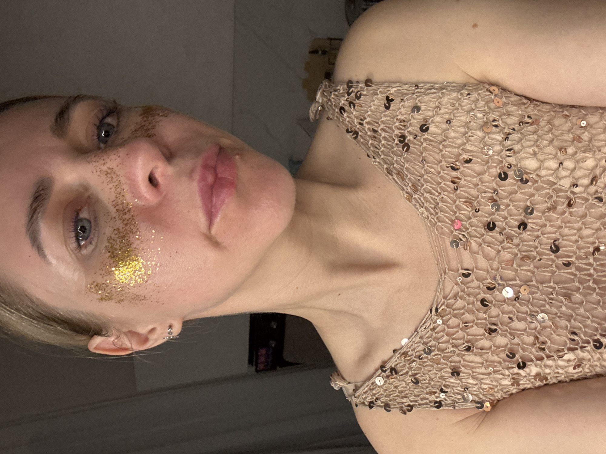 A close-up, vertically oriented portrait features a young adult female, positioned with her head tilted to her right. She has light skin, blue-grey eyes, and light-colored hair, partially visible around her ear. Her lips are slightly parted, revealing a soft pink tone.
A prominent detail is the application of chunky gold glitter on her face, concentrated on her right cheekbone, extending under her right eye and towards her nose, indicating a festive or artistic makeup style. She is wearing a light beige or nude-colored top, made of a loosely woven or crocheted fabric, embellished with numerous small, round sequins in varying metallic shades including bronze, silver, and rose gold. A small silver-colored hoop earring is visible in her right earlobe.
The subject is illuminated by soft, directional lighting that highlights the textures of her skin, the glitter, and the sequins on her garment. The background is dark and out of focus, suggesting an indoor setting with subdued lighting. Her expression is neutral, and her gaze is directed towards the viewer. No visible text is present in the image.