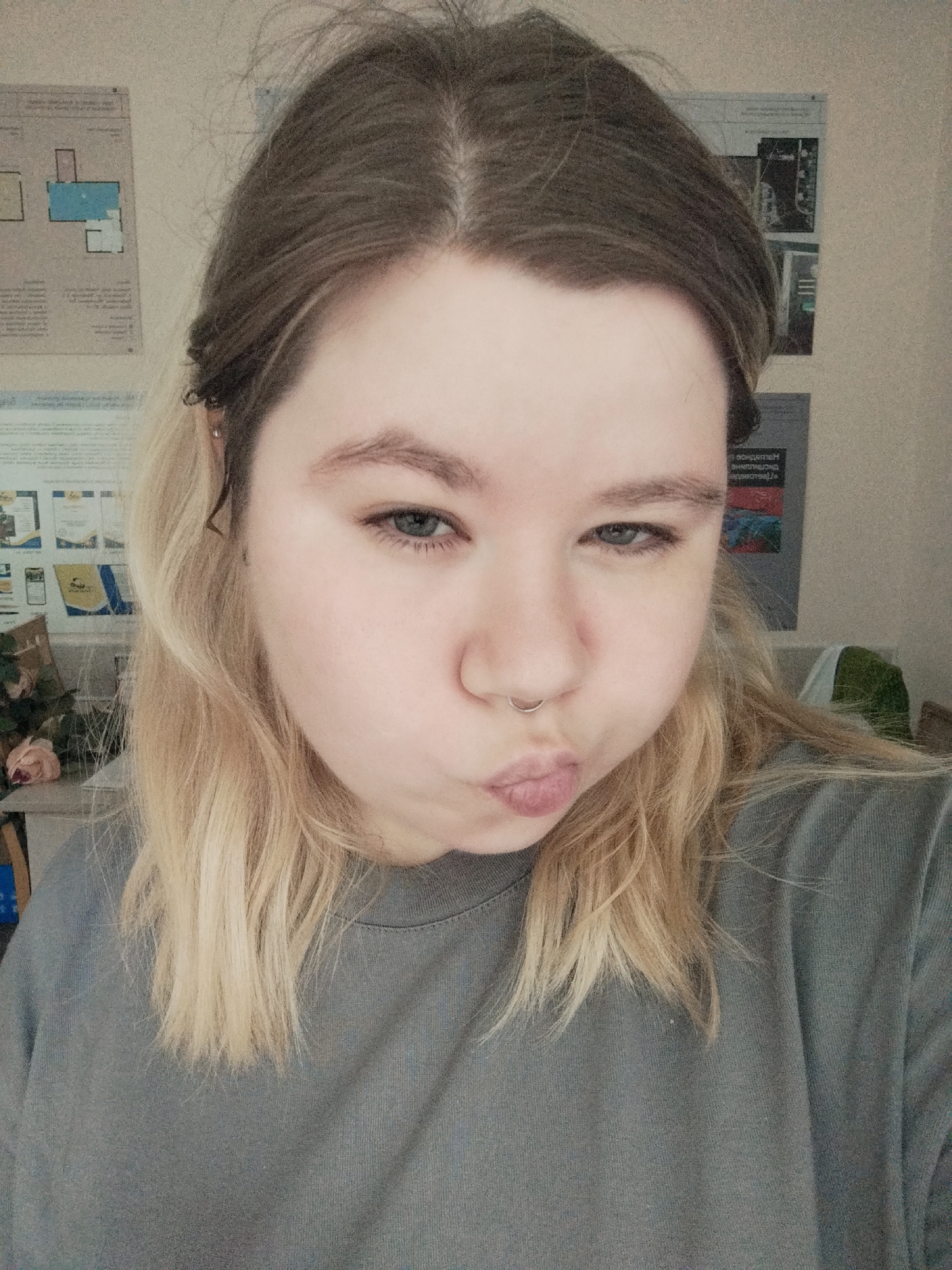 A young woman with blonde hair pulled back from her face and a septum piercing is shown from the chest up in a selfie. She is looking at the camera with her lips pursed. She is wearing a gray sweatshirt. In the background, posters and a bouquet of flowers are visible. The lighting is soft and diffused.