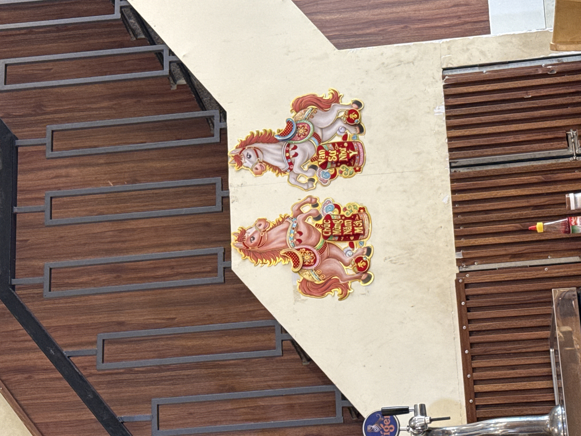 The image depicts a decorative display of two stylized horses with gold trim, positioned on a cream-colored wall adjacent to a wooden staircase. The horse on the left is predominantly pink and brown, adorned with a saddle and a sign that reads "Chúc Mừng Năm Mới" (Happy New Year). The horse on the right is white and reddish-brown, with a similar saddle and a sign that says "Vạn Sự Như Ý" (May all your wishes come true). Both horses are decorated with floral and cloud motifs. The background features a wooden paneled staircase with dark gray metal railings. To the left, a chrome beer tap with a blue "Tiger" logo is visible, suggesting a bar or restaurant setting. Below the wall, a dark brown wooden slatted structure is present, and a bottle of red sauce with a yellow cap is seen on the right. The scene is indoors, likely in Nha Trang, Vietnam, given the Vietnamese inscriptions on the horse decorations. There are no people visible in the image.
