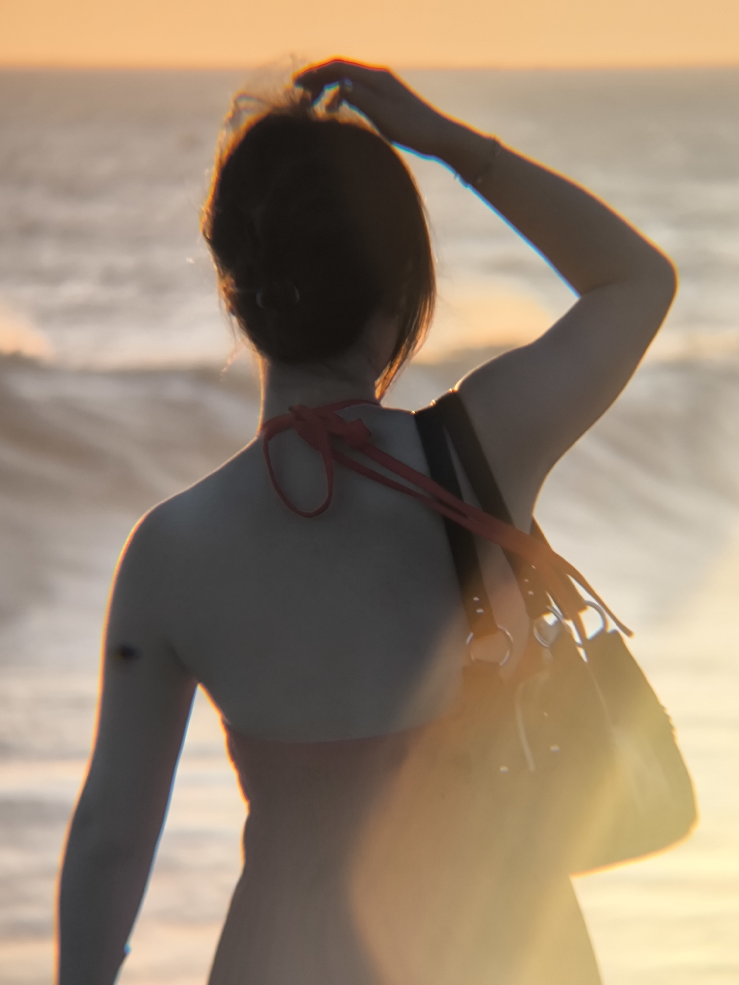 A woman stands on a beach, looking out at the ocean. She is wearing a red swimsuit and carrying a black bag over her shoulder. Her hair is pulled back. She has her right hand on her head. The sun is setting in the background.