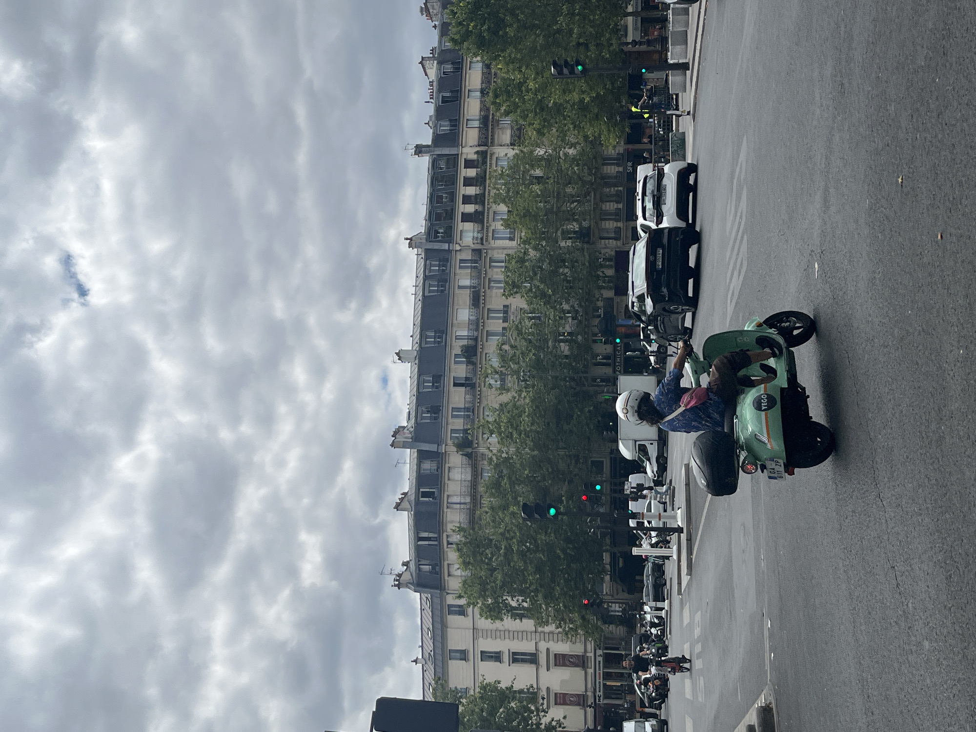 An eye-level medium shot captures a street scene in Paris, France, on a cloudy day. In the foreground, a person rides a light green electric scooter with the brand "Yego" visible on its side. The rider, wearing a white helmet and a blue patterned shirt with a pink bag slung across their chest, is turned away from the camera, looking towards the right. The scooter has a black storage box on the back. The road is asphalt, with white markings indicating a bus lane.
In the mid-ground, several cars are stopped at a traffic light, which is green for the direction the scooter is facing. A black SUV and a white compact car are prominent. Further back, a white delivery van and various other vehicles are visible, along with other people on motorcycles and scooters.
The background is dominated by a multi-story building with a mansard roof, characteristic of Parisian architecture. Numerous trees line the street in front of the building, their green foliage contrasting with the overcast sky. Traffic lights and streetlights are also present. The overall atmosphere is that of a busy urban intersection.