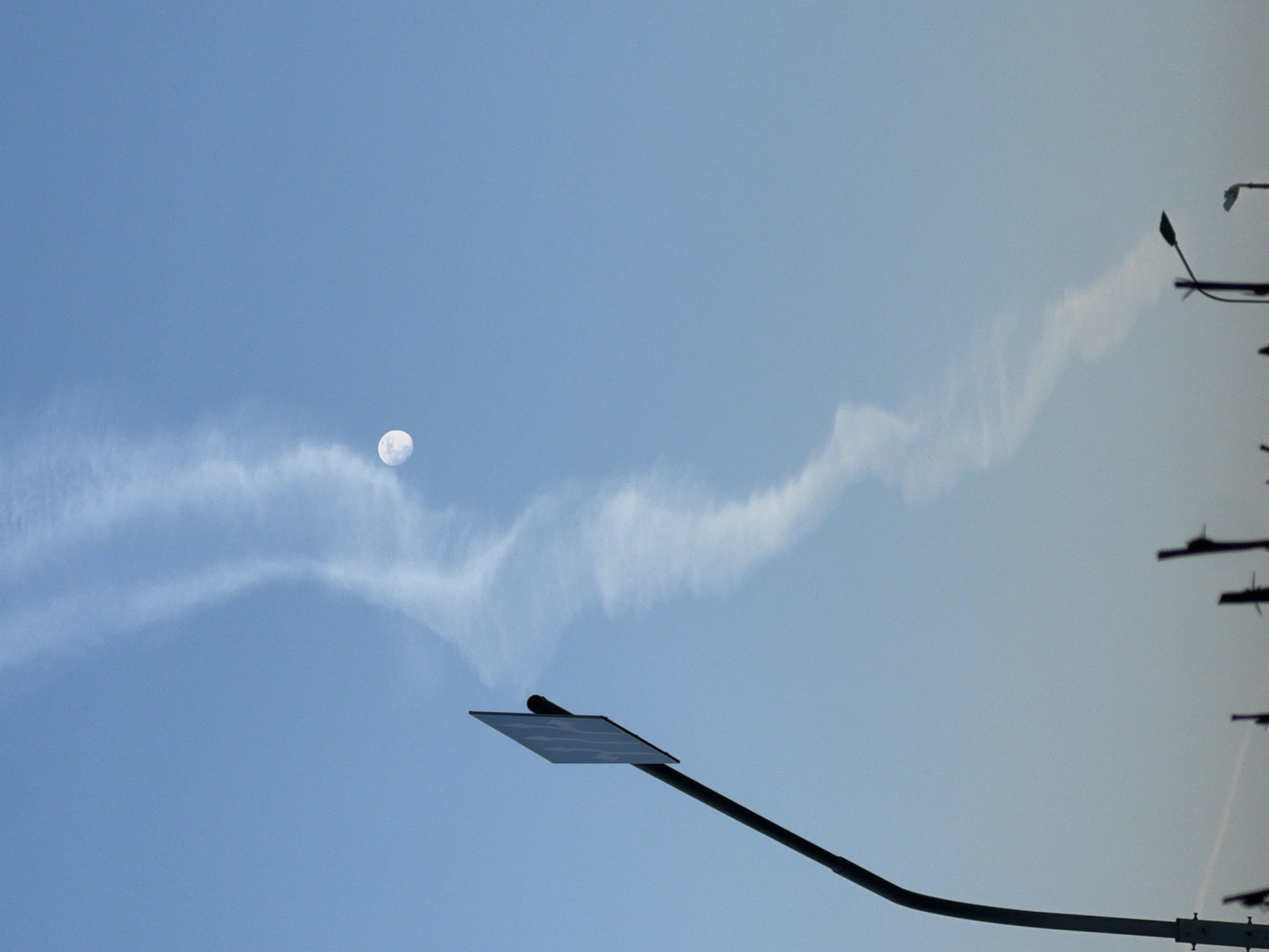 The image captures a daytime sky in Saint Petersburg, Russia. The sky is predominantly blue, with wispy, cloud-like formations stretching across the frame. The moon is visible as a pale, circular object within the cloud formations. In the foreground, a street lamp with a rectangular head is positioned. Further to the right, silhouetted against the sky, are utility poles and wires.