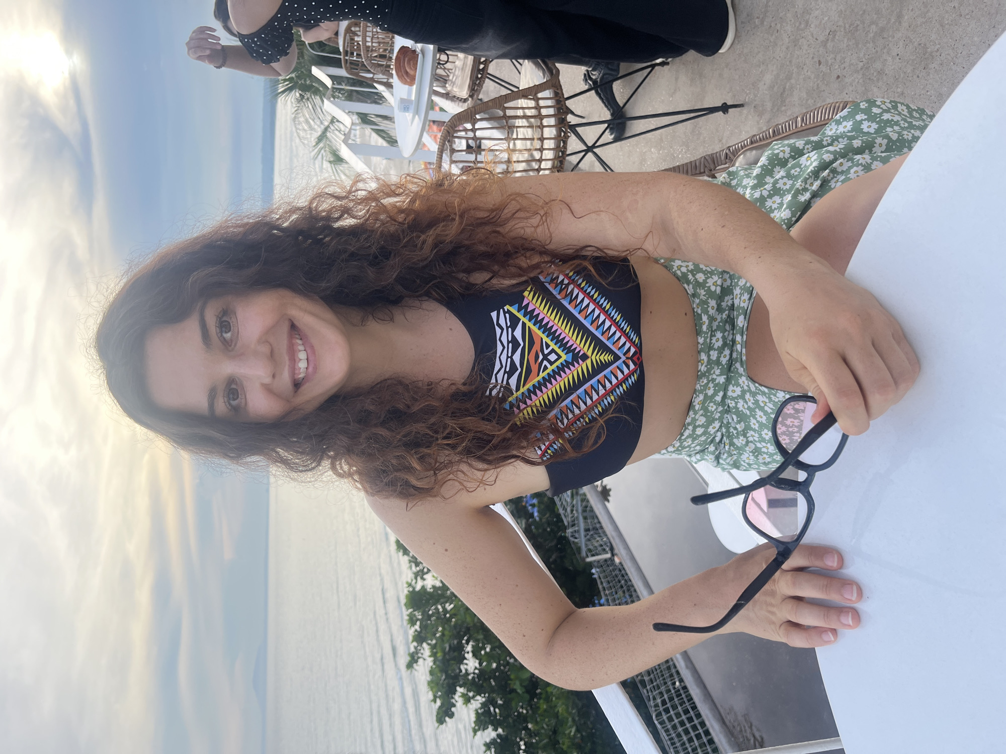 A woman with long, curly, auburn hair is seated at a white table outdoors, holding a pair of black-rimmed glasses. She is wearing a black bikini top with a colorful, geometric pattern and a light green skirt with a white daisy print. She is smiling and looking at the camera. Behind her, the ocean stretches to the horizon under a cloudy, pale sky. To her right, another person, partially visible, is wearing a black top with white polka dots. The setting appears to be a restaurant or cafe with wicker chairs and a white railing visible in the background. The image was taken in Mũi Né, Vietnam.