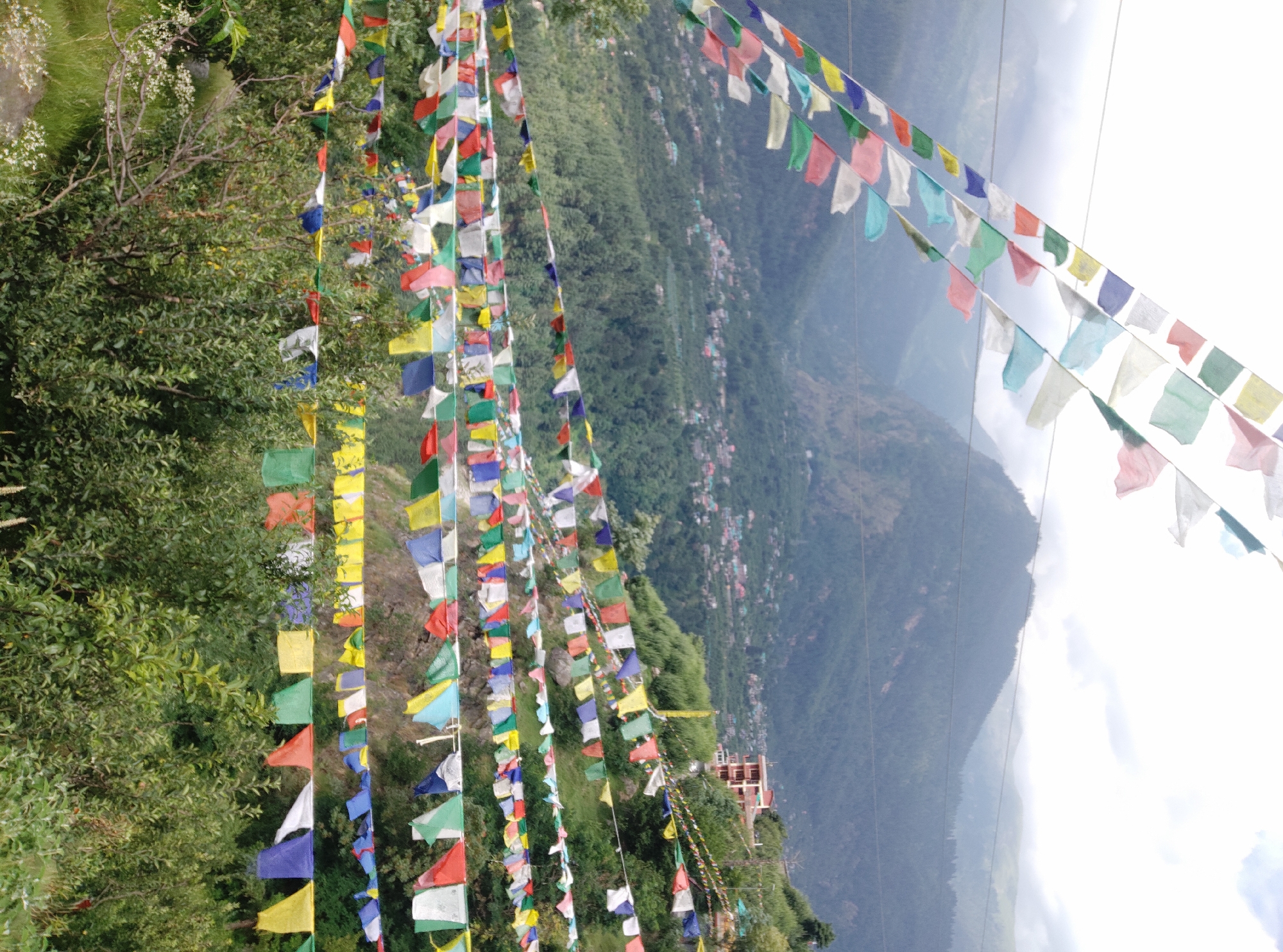 The image depicts a scenic mountain landscape in Baragran, India, adorned with numerous strings of colorful prayer flags. The flags, in shades of blue, yellow, red, green, and white, are strung across the foreground, fluttering in the breeze. They are set against a backdrop of lush green hills and a hazy sky, suggesting a mountainous region. In the mid-ground, a village or town is visible nestled on the slopes of the mountains, with buildings appearing in various colors. The time of day appears to be daytime, with natural light illuminating the scene, though the sky's brightness suggests it could be late morning or early afternoon. The weather seems mild, indicated by the clear visibility of the landscape and the gentle movement of the flags. The overall impression is one of tranquility and spiritual significance, often associated with prayer flags. There is no visible text in the image.