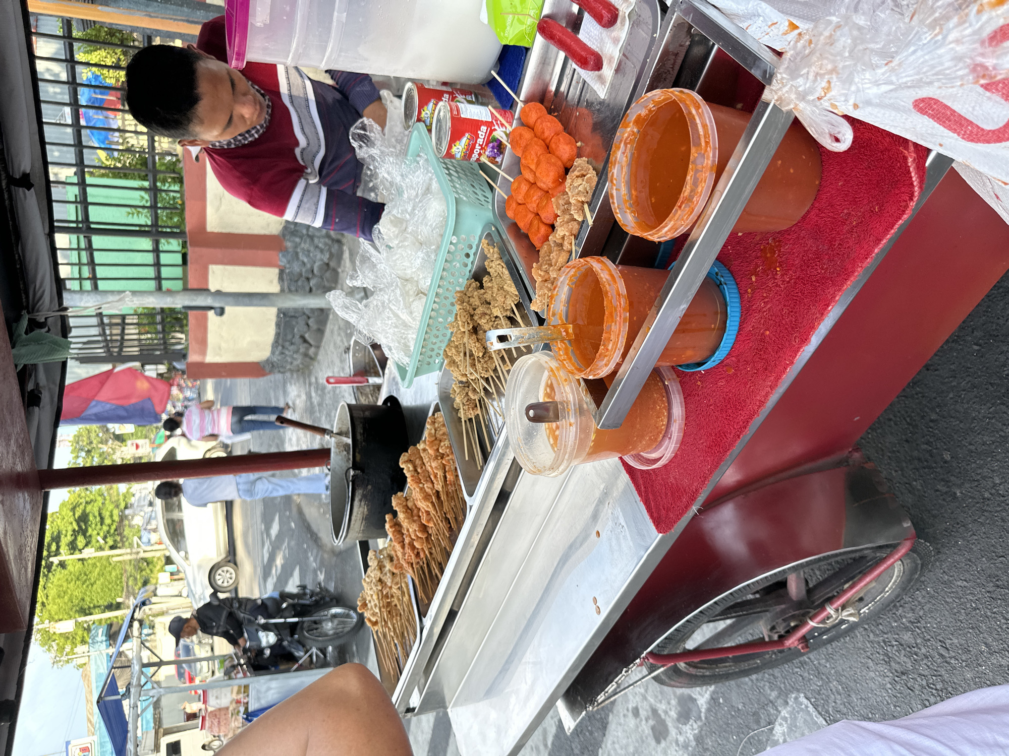 A street vendor in Davao City, Philippines, is preparing food at a mobile cart. The cart is equipped with various food items, including skewers of fried meat, fish balls, and sausages. There are also large containers of dipping sauce and a basket of plastic bags. The vendor is wearing a red and white striped shirt and is in the process of serving a customer. In the background, there are cars and motorcycles, indicating this is a busy street. The scene suggests a typical street food setup in the Philippines, with fresh ingredients and a lively atmosphere.