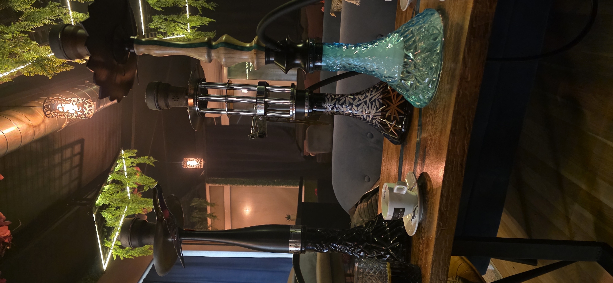 Three hookahs are placed on a wooden table in what appears to be a dimly lit establishment, potentially a café or lounge. The first hookah, closest to the foreground, has a black stem and a decorative black and silver base. Next to it is a white cup and saucer. The second hookah features a silver stem and a geometric-patterned black and bronze base. The third hookah has a wooden stem with green accents and a bright blue, crystalline-patterned base. In the background, there is a couch, and the room is decorated with hanging lights and greenery. The location is likely in Sofia, Bulgaria, given the architecture and interior design elements.