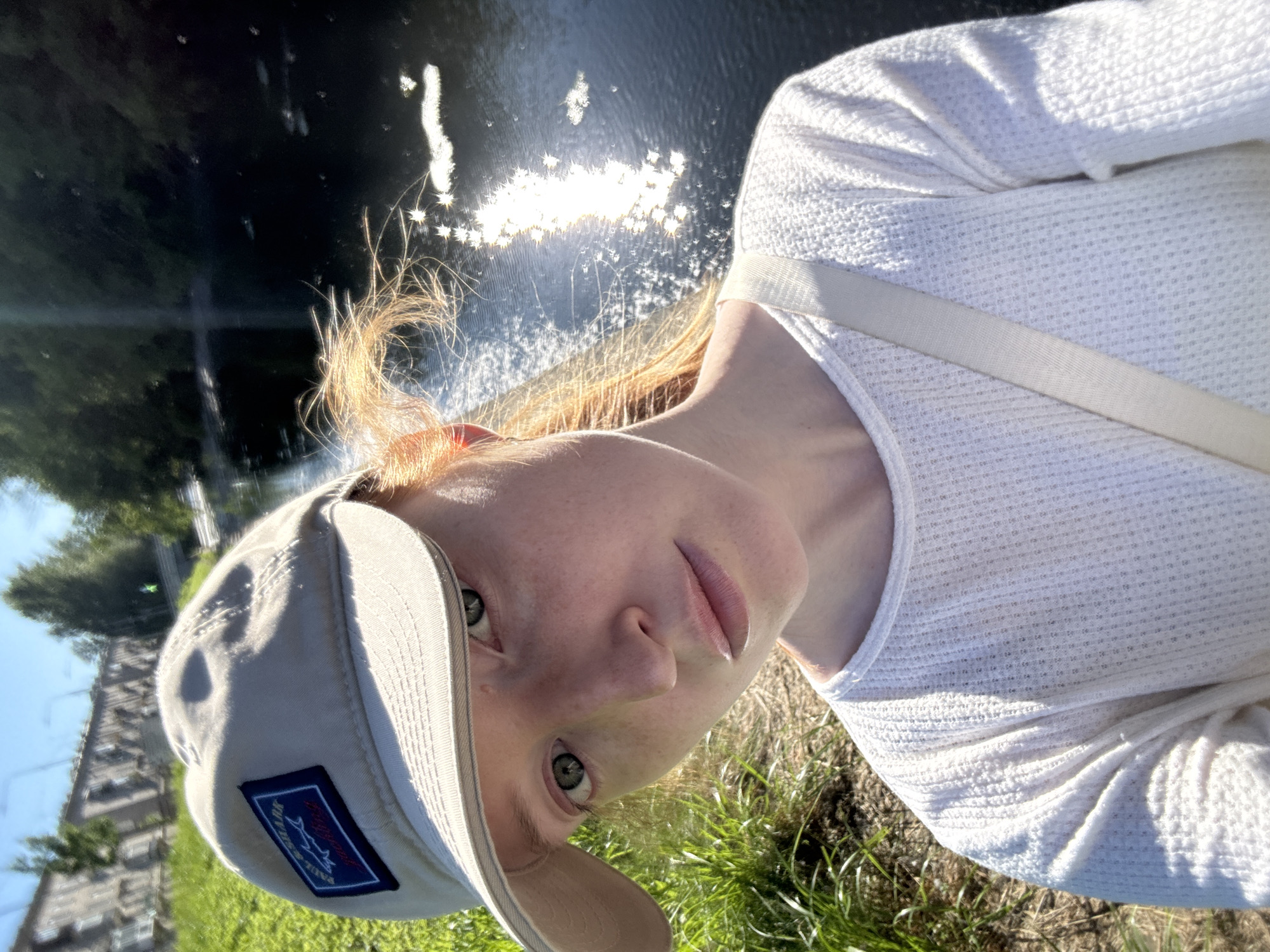 A young woman with red hair, wearing a light-colored baseball cap and a white long-sleeved shirt, is pictured outdoors on a sunny day. She is positioned in the foreground, looking towards the camera with a neutral expression. Her hair is partially visible, appearing windblown. A beige strap is visible across her shoulder.
Behind her, a dark body of water reflects the bright sunlight, creating a shimmering effect on its surface. Lush green grass lines the bank of the water. In the background, there are trees and a glimpse of a building structure, possibly industrial or urban, with a bridge visible in the distance. The overall impression is of a candid outdoor portrait taken near a waterway within a city environment. The presence of a building and potential urban infrastructure suggests a location within Saint Petersburg, Russia, likely near a park or a waterfront area like the Neva River or one of its canals.