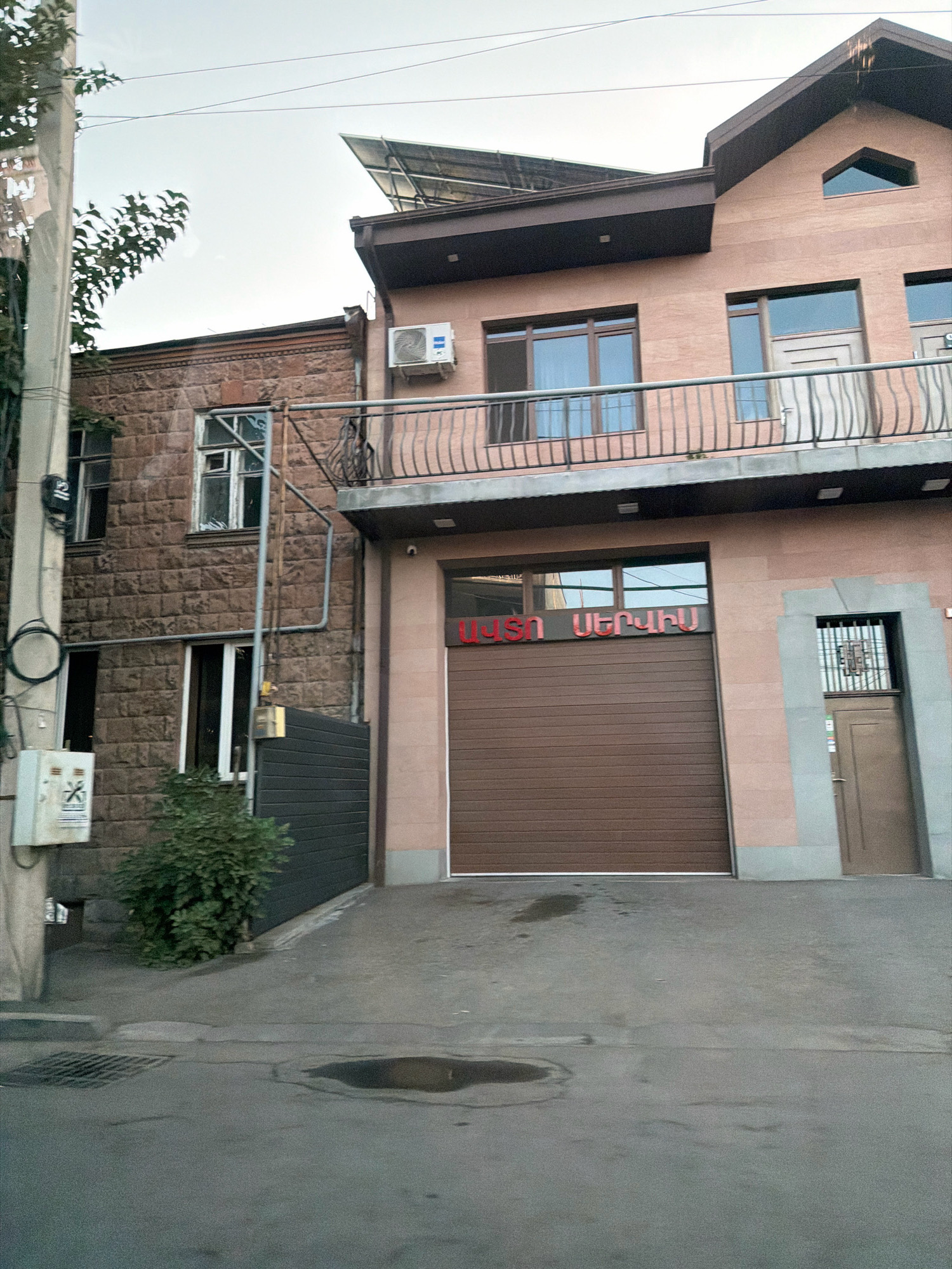 The image depicts a section of a street in Yerevan, Armenia, featuring a two-story building with a brown garage door and a balcony. The building's facade is a mix of stone and painted surfaces. Above the garage door, red Armenian lettering is visible on a horizontal strip. To the left of the garage is a stone-clad section of the building with several windows. A utility pole with some electrical boxes and wires stands in front of this section. To the right of the garage, there is a smaller door with a metal grate over a window. The ground in front of the building is paved with asphalt, and there are a few puddles of water on the surface. Lush green bushes are planted to the left of the garage, against the stone wall. The sky above is overcast. The building appears to be a residential or commercial property, possibly a shop or workshop with living quarters above. The overall impression is of a functional urban environment.