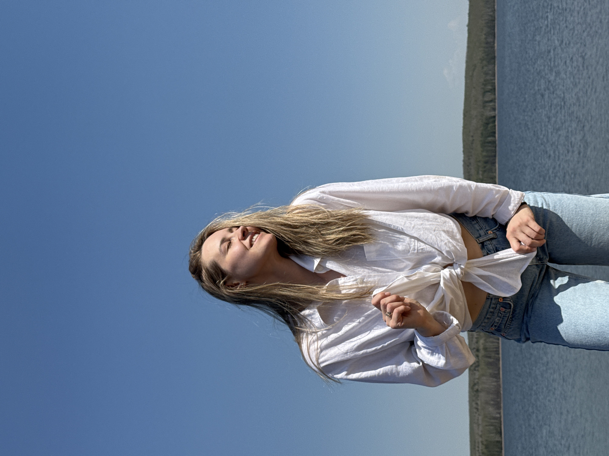 A young woman is photographed outside on a sunny day in Chaikovsky, Russia, adjacent to the Kama River. She's wearing light blue jeans and a white, long-sleeved shirt tied at the waist, exposing a section of her midriff. Her head is tilted back, her eyes are closed, and she is smiling. The blue sky fills the upper portion of the image. To the right, a body of water, identified as the Kama River, extends into the background and is separated from the sky by a strip of greenery on the opposite riverbank.