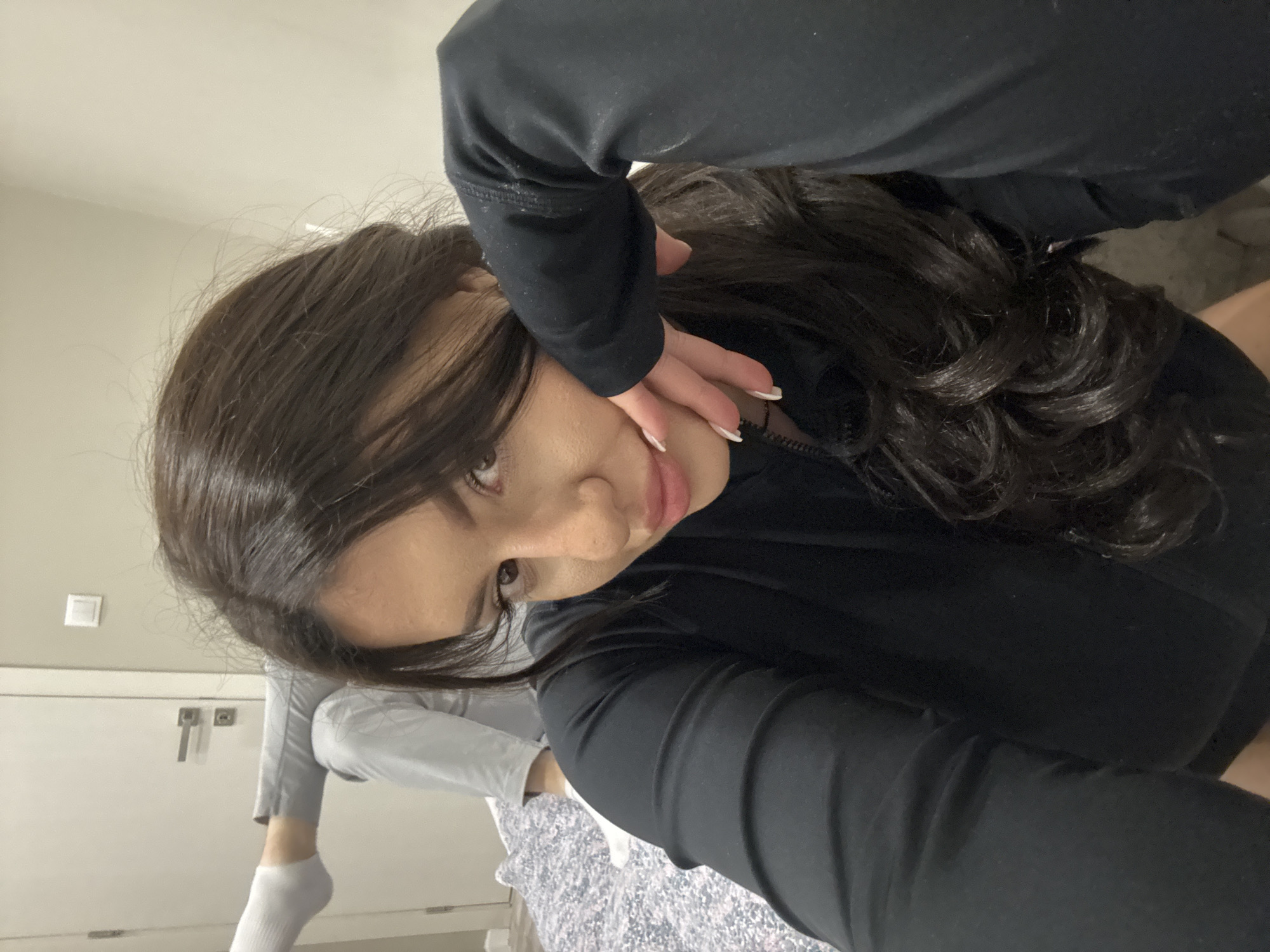 A woman with long, dark hair and fair skin is posing for a selfie. She is wearing a black long-sleeved top. Her left hand is touching her face near her eye. The background is a bedroom with a white door and cabinet. A person in gray pants and a white sock is visible in the lower-left corner of the image. The setting seems to be indoors, on a patterned bedspread. There is no information in the image that definitively places the location in Burgas, Bulgaria.