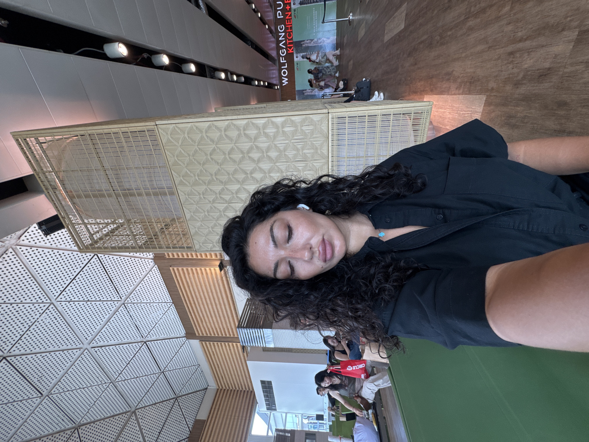 The image, correctly oriented, is a selfie capturing a woman with long, dark curly hair, wearing a black collared shirt. She has light-colored earbuds in her ears and a small turquoise pendant necklace. Her eyes are closed, and she appears to be resting or relaxed. Her arm and shoulder are visible in the foreground, indicating she is holding the camera.
The background reveals a spacious, modern indoor environment, likely a public or commercial space such as an airport terminal or a large food court in Kuta, Indonesia. Architectural elements include a wall or ceiling with patterned triangular, perforated panels on the left, and further back, walls with horizontal wooden slats. A tall, light-colored lattice-work structure, possibly a decorative screen or partition, stands prominently in the mid-ground.
Several other people are visible in the background, sitting or waiting. One person has a red bag with the word "CIRCLE" visible, and a black backpack and white sneakers are seen next to another individual. Overhead track lighting with multiple spotlights illuminates the area, suggesting daytime. A large vertical sign in the far background displays partial text: "WOLFGANG PU" and "KITCHEN + B", which likely refers to a "Wolfgang Puck Kitchen + Bar" establishment. The overall scene depicts a moment of calm within a potentially busy setting.