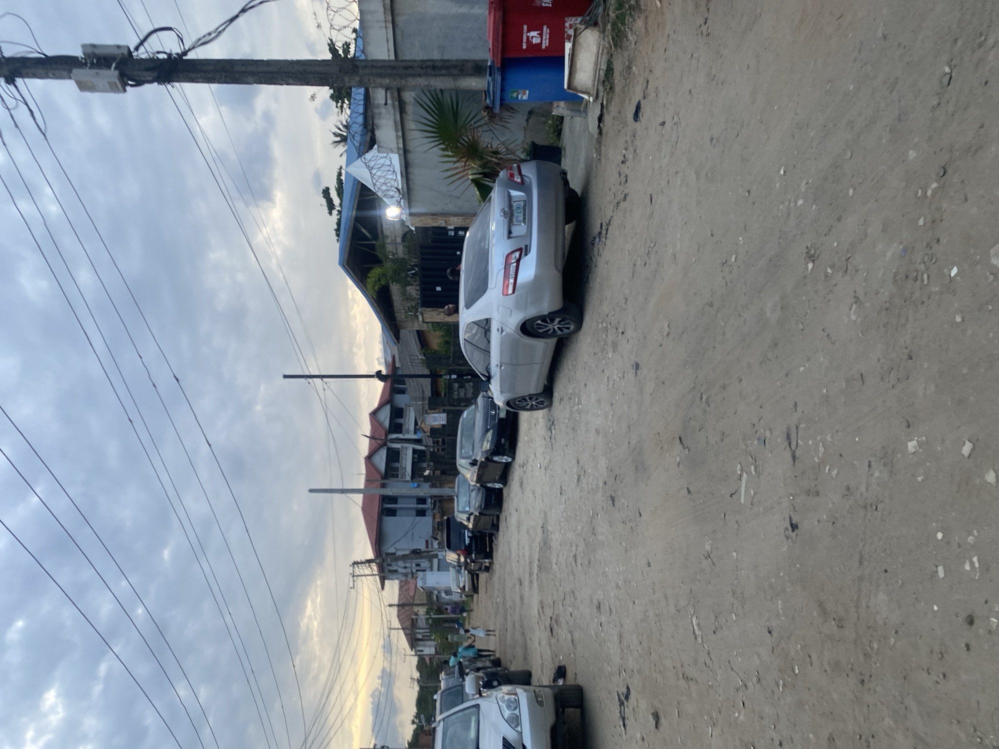 The image depicts a street scene in Lagos, Nigeria. A row of parked cars lines the left side of a dirt road, with buildings visible in the background. Utility poles with wires run along the left edge of the frame. A red and blue trash can is positioned near the beginning of the row of cars. The sky is overcast with cloudy conditions. There are people in the distance.