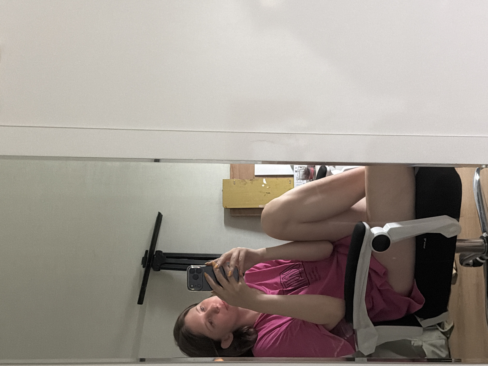 A young woman is captured in a mirror selfie, seated in a white and black office chair. She is wearing a bright pink t-shirt and is holding a smartphone in her hands, angled to take the photo. Her legs are crossed, and her posture suggests a casual, relaxed demeanor. The background is a lightly colored wall, with what appears to be a portion of a white cabinet or door to the right of the mirror. To the left of the woman, a black metal stand is visible. The environment suggests an indoor setting, possibly a home or studio. The specific location within Dongducheon-si, South Korea cannot be determined from the image alone.