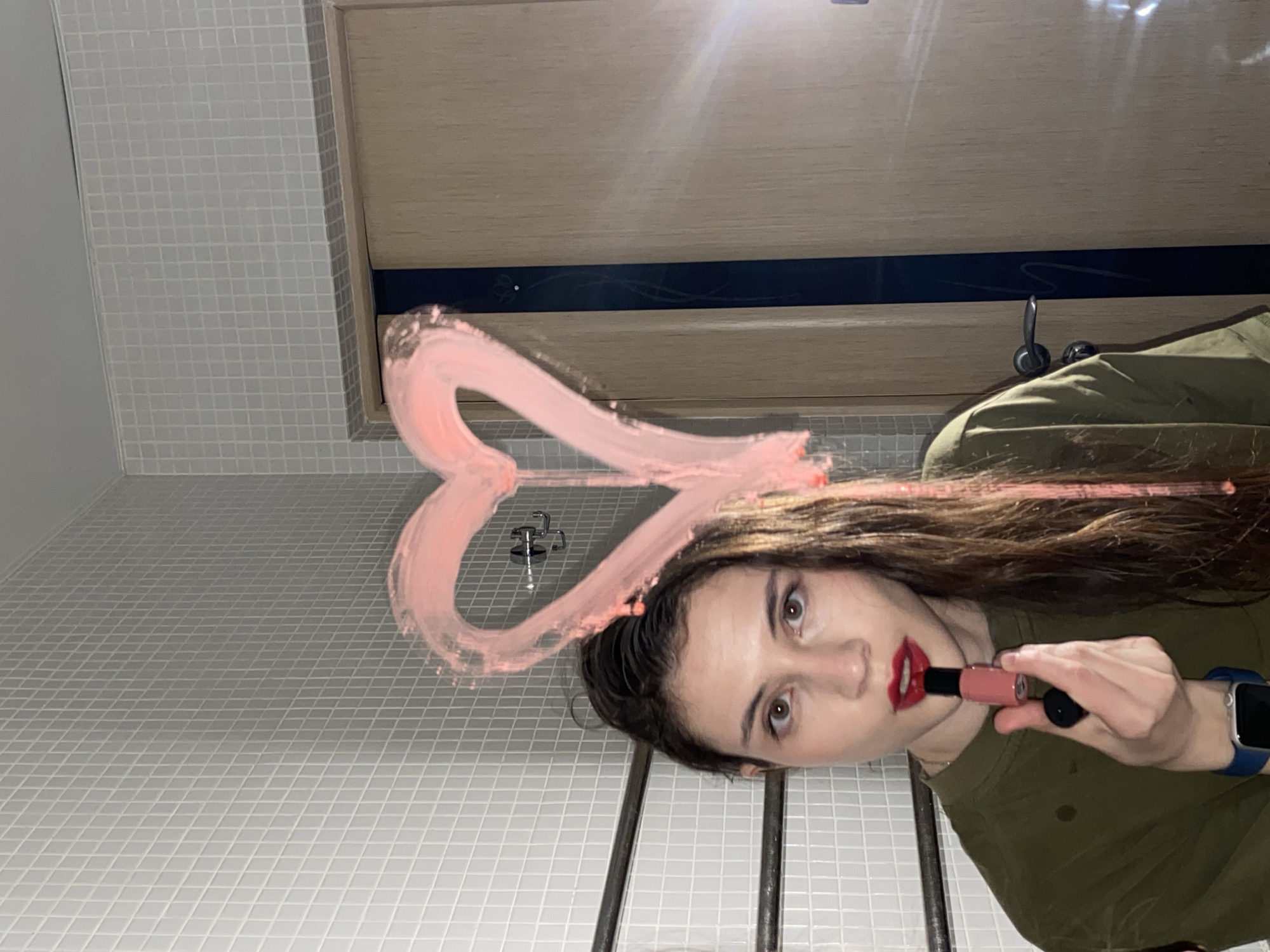 The image is a low-angle, rotated selfie taken indoors, possibly in a bathroom, looking upwards at a young woman. She has long, dark brown hair, with strands near her face appearing pink, possibly stained by lipstick. She wears a dark green top and a smartwatch with a blue band on her left wrist. Her lips are covered in red lipstick, and she holds an open tube of lighter pink liquid lipstick in her right hand, positioned near her mouth as if applying it.
Above and to her left, a large, pink heart shape is drawn with lipstick on a reflective surface, likely a mirror. The background consists of light-colored, small square tiles on the walls. Above the tiled section, there's a wooden or laminate panel featuring a black horizontal strip with a subtle, decorative floral or swirl pattern. A silver faucet or tap fixture is visible in the upper left background. Bright artificial light sources create glare in the upper part of the image. The woman's expression is focused and serious as she looks directly towards the camera. The setting is Votkinsk, Russia. No visible text is present in the image.