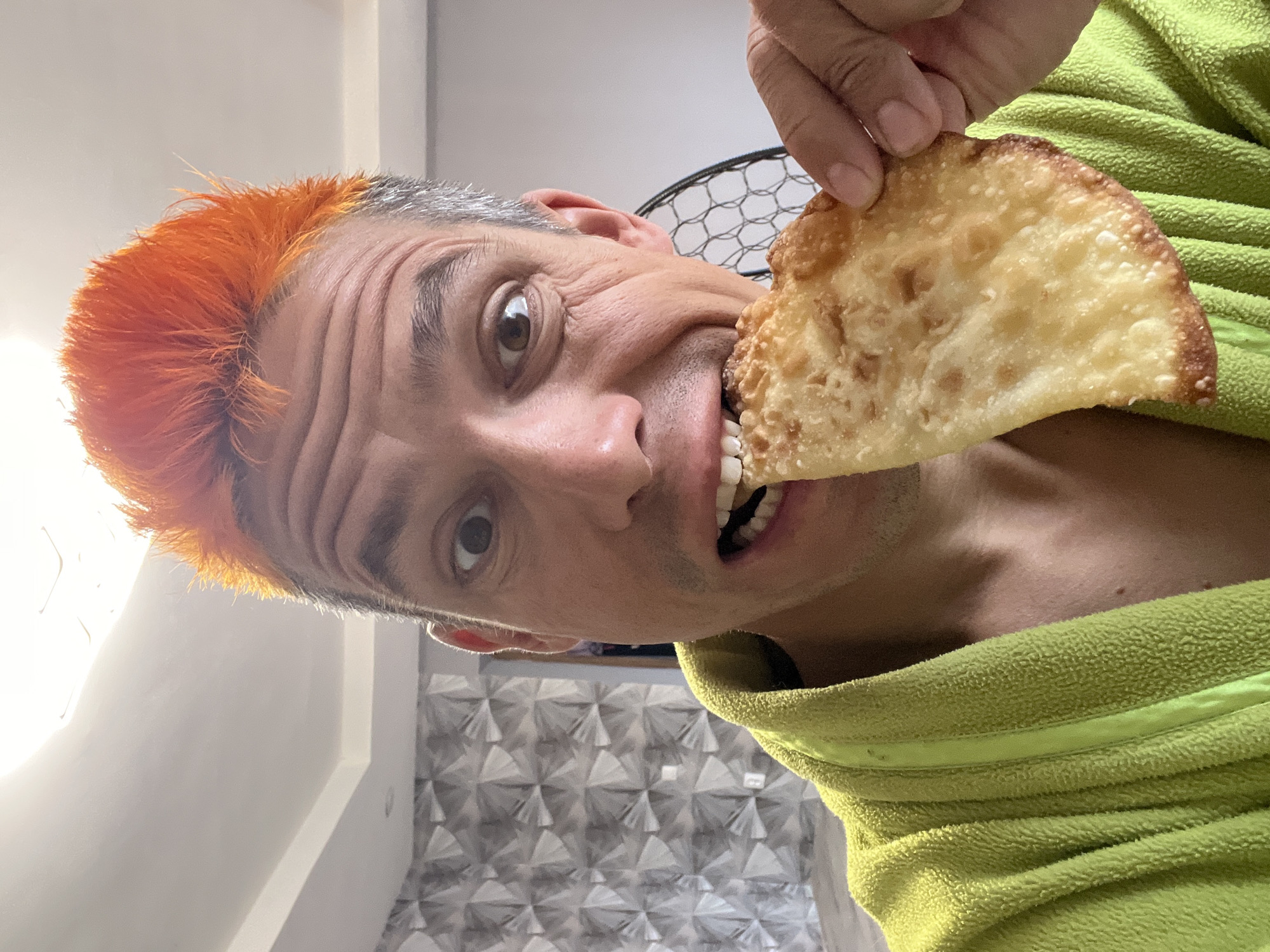 A close-up, tilted photograph captures a man with brightly colored orange hair on top and grey at the sides. He is in the process of eating a piece of fried dough or flatbread, which he is holding with his fingers. The man is wearing a green, fleece-like garment. The background includes a white wall and a geometric patterned wall covering. The photograph was taken indoors in Chaikovsky, Russia.