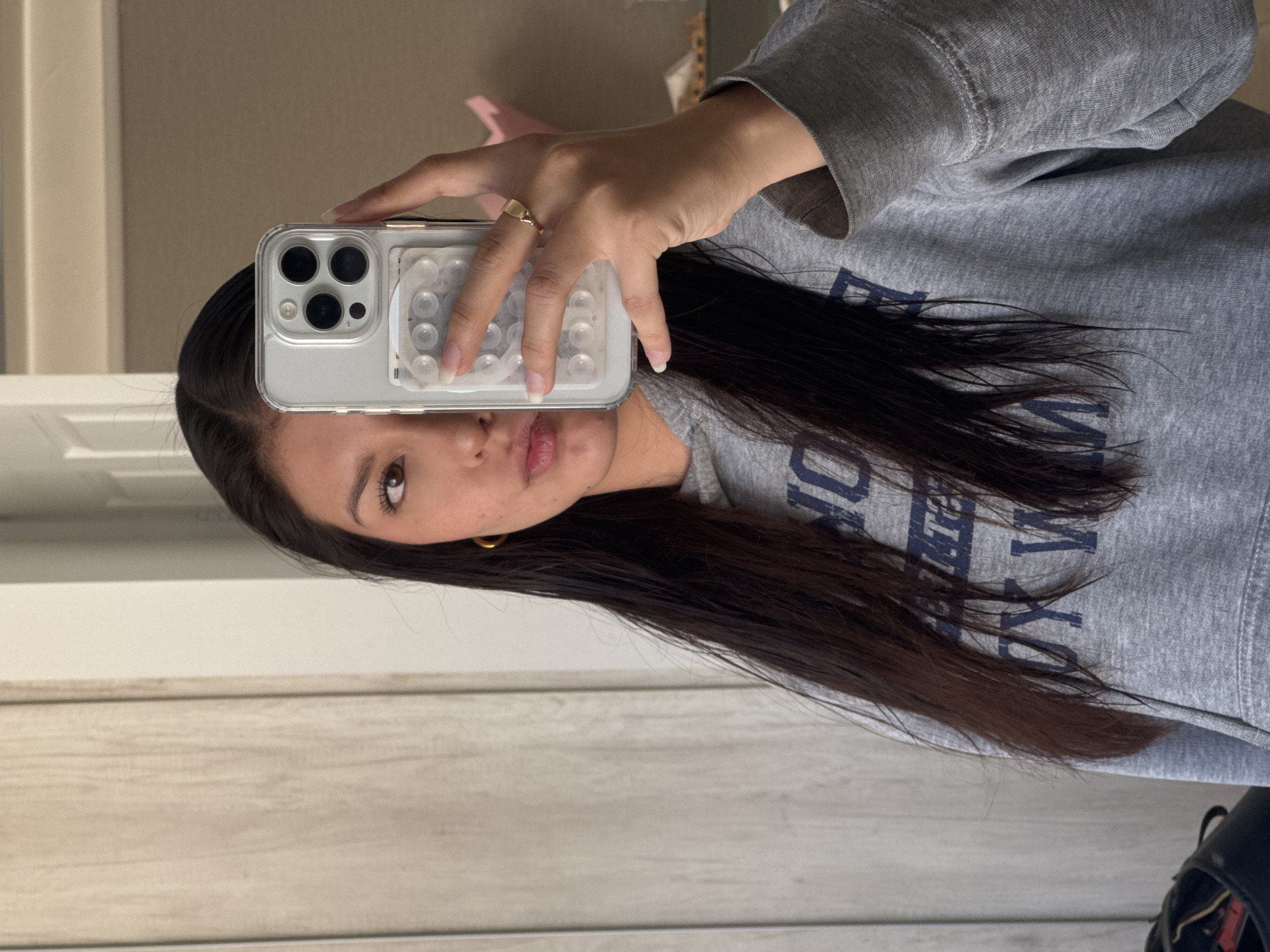 A young woman is holding a phone in front of her face, apparently taking a selfie. She is wearing a grey sweatshirt with dark blue lettering on it. She has long, dark hair and is wearing a gold ring on her finger. The phone case is clear and has a grid of white circles on the back. The background is slightly out of focus, but appears to be a room with light-colored walls and trim. There is no indication of Ocotlán, Mexico, or San Francisco in the image.