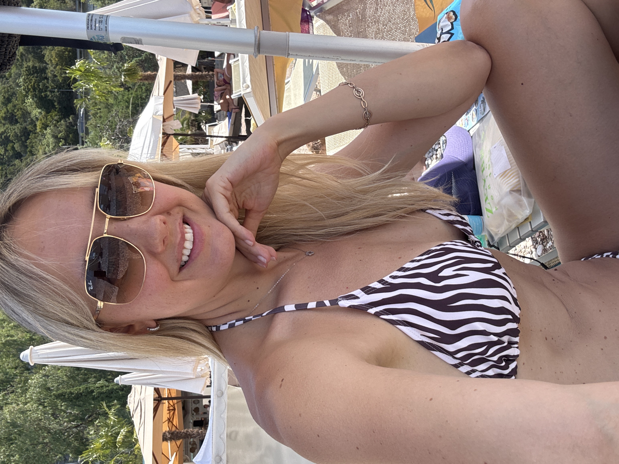 A blonde woman wearing a zebra-print bikini and sunglasses is pictured at the Kamenovo beach in Montenegro. She is seated outdoors, with her right arm bent and hand resting on her chin, and her left arm resting on her leg. In the background, beach umbrellas and lounge chairs are visible, as well as trees and hills. The lighting suggests it is daytime and the weather is sunny.