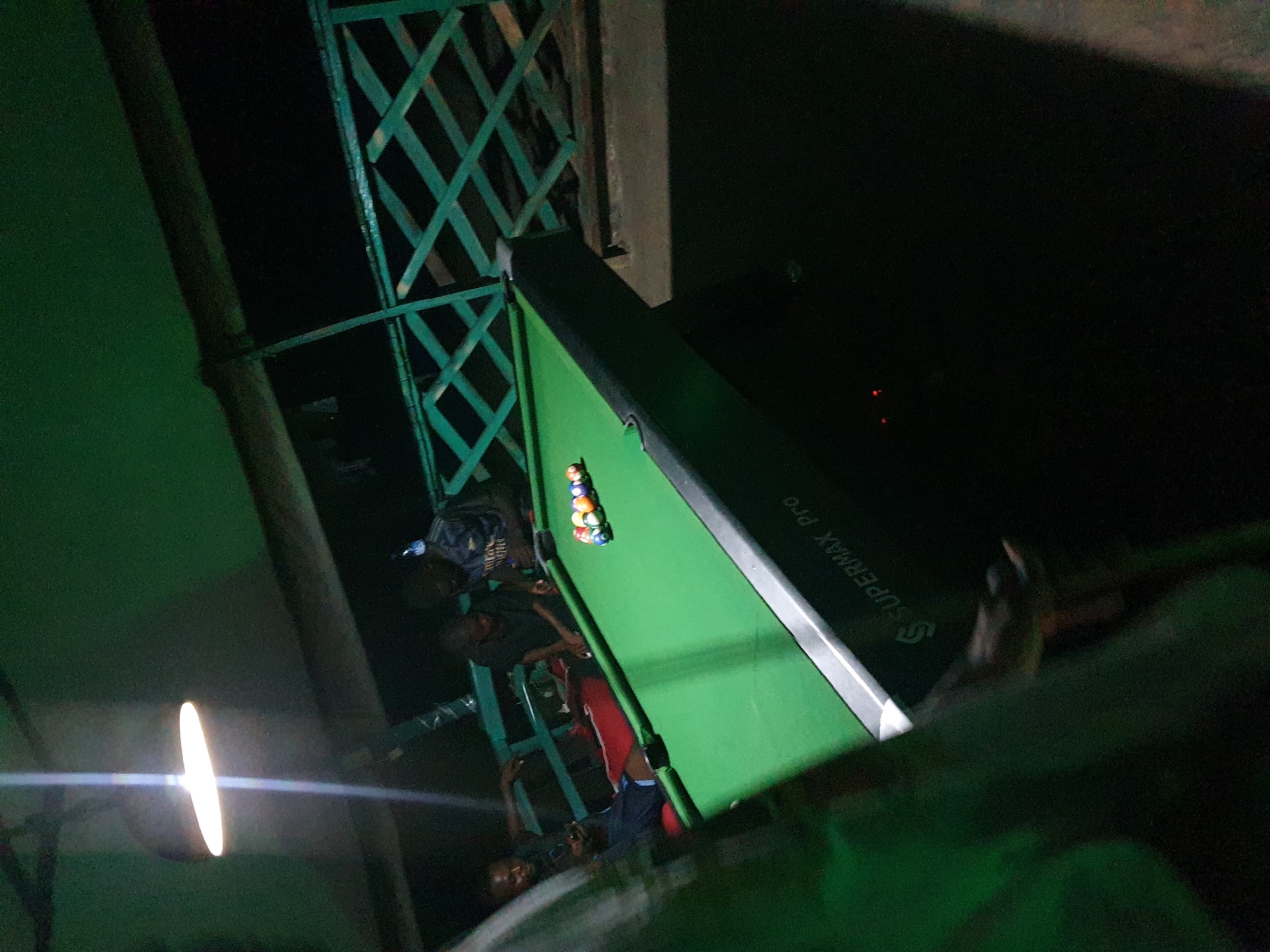The image depicts a dimly lit outdoor scene, likely a rooftop or balcony in Lokoja, Nigeria, where a group of young men are gathered around a green pool table. A bright, overhead light illuminates the pool table and the immediate surroundings. The pool table is set up with a rack of colorful billiard balls at one end. Three individuals are visible near the table; one is seated and looking at his phone, another is standing and appears to be observing the game, and a third is seated behind the table, his face obscured. The background shows a dark environment with hints of a green metal railing or fence, suggesting an elevated or enclosed outdoor space. The overall atmosphere appears casual and social, with the group engaged in recreational activity at night. The "SUPERMAX Pro" branding is visible on the side of the pool table.