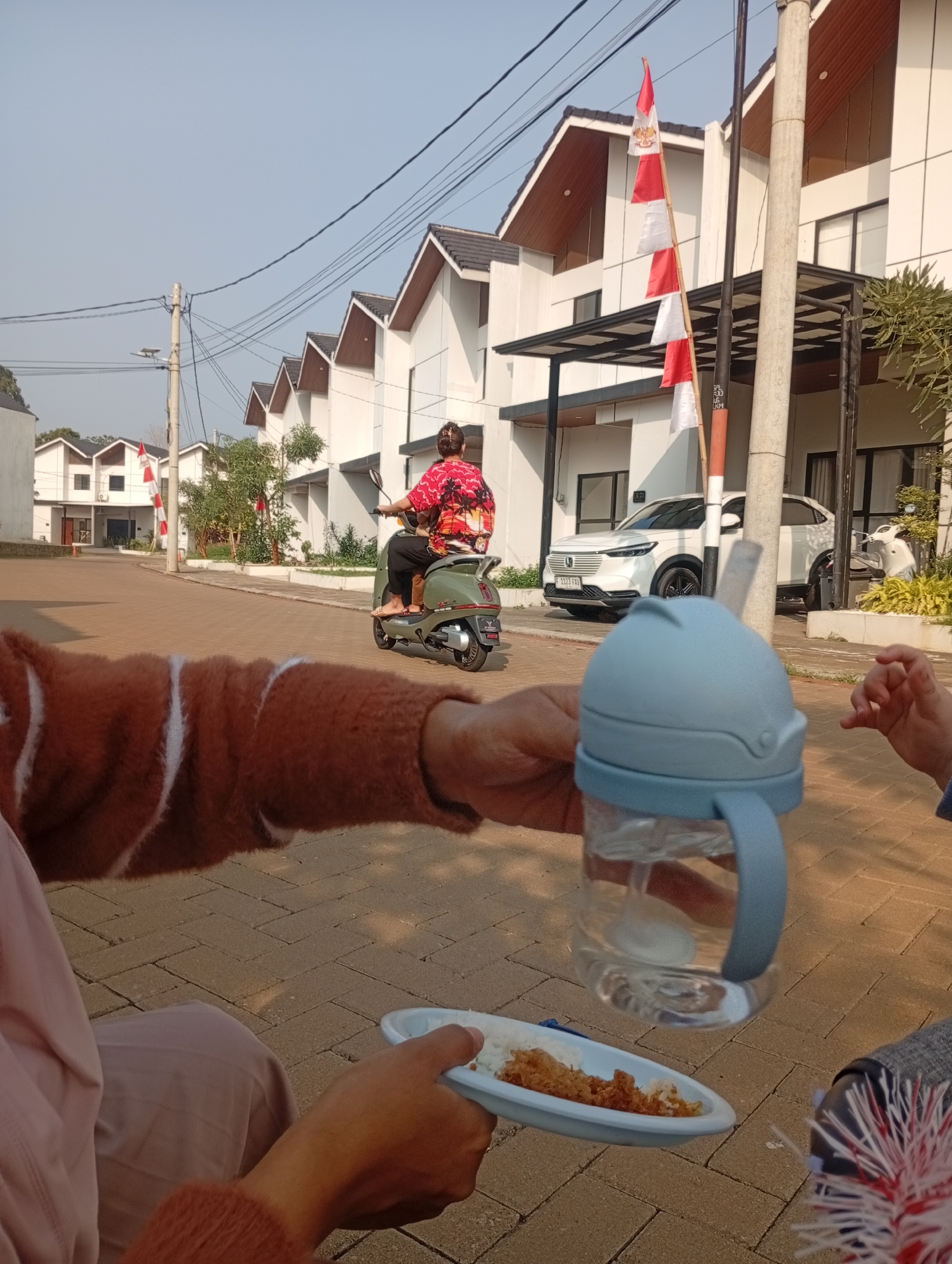 The image captures a street scene in Kemang, Indonesia, characterized by modern residential buildings with white facades and dark roofs, lined with palm trees. The foreground is dominated by a person's arm, wearing a brown sweater, holding a light blue sippy cup filled with water and a straw. Another hand in the foreground holds a small white plate with a serving of rice and shredded chicken. In the mid-ground, a woman on a green scooter, wearing a vibrantly patterned red and pink shirt, drives past. Beside her, a white SUV is parked in front of one of the houses. A red and white Indonesian flag is prominently displayed on a pole near the street, indicating a possible national celebration or commemoration. The street is paved with interlocking concrete blocks, and the sky is clear and blue, suggesting a sunny day. The overall ambiance is one of daily life unfolding in a suburban Indonesian neighborhood.