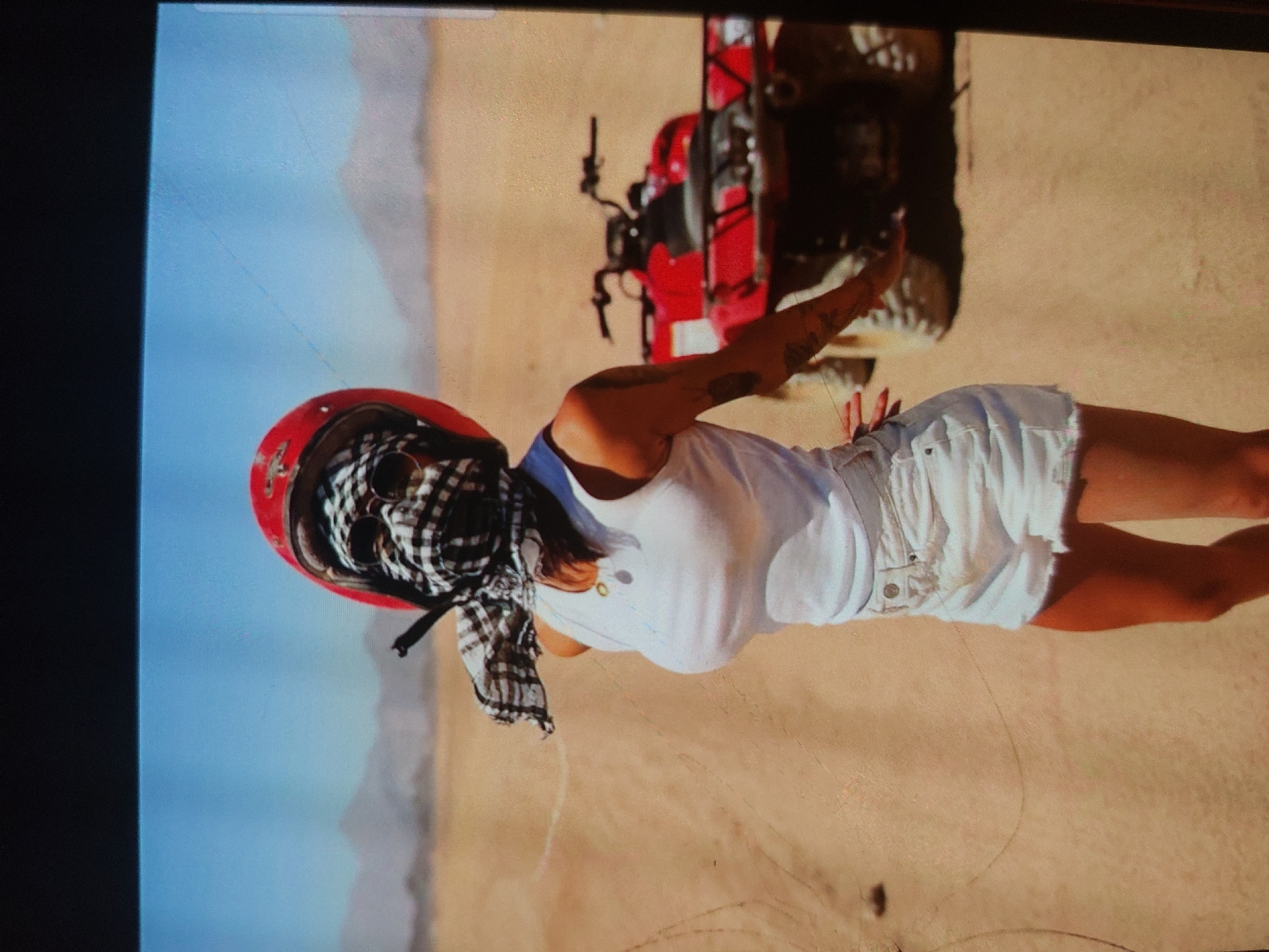 A full-body shot depicts a person, likely a woman, standing upright in a desert or sandy landscape next to a red quad bike. She is positioned towards the right of the frame, looking slightly left. She wears a red full-face helmet, with a black and white checkered keffiyeh-style scarf covering her lower face. Her attire includes a white tank top and light-wash denim shorts. Tattoos are visible on both arms.
Behind her, partially visible, is a red quad bike with black components. The setting is an arid outdoor environment, characterized by expansive light brown, sandy terrain with faint tracks, and distant, muted hills or mountains under a clear, light blue sky. The lighting suggests it is daytime, likely sunny, casting a shadow from the person onto the sand. The overall scene conveys an activity such as quad biking in a recreational desert setting. No text is visible in the image. The location context provided is Tsarevo, Bulgaria.