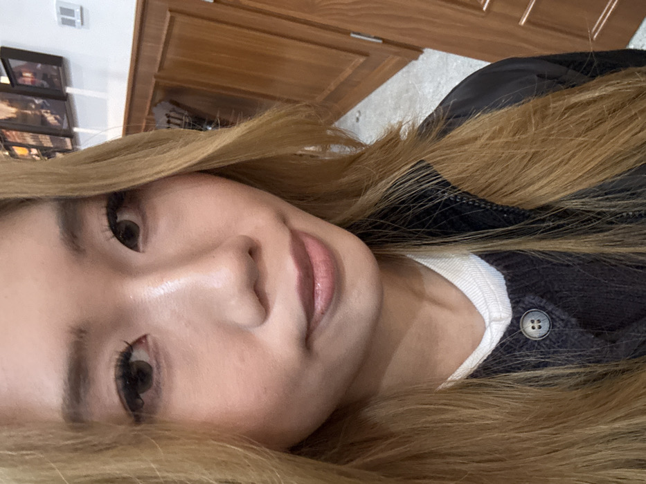 A close-up, waist-up portrait captures a female subject, appearing to be East Asian, looking directly at the camera. Her long, light brown hair cascades over her shoulders and chest. She has fair skin, prominent eyelashes, dark eyes, and her lips are slightly parted in a neutral expression. She wears a white top beneath a dark, possibly charcoal or black, ribbed cardigan or sweater with a visible gray button. A dark, shiny material, likely a jacket or vest, is visible over her right shoulder. The background indicates an indoor environment, featuring a two-toned wall with light brown wooden paneling on the lower section and a plain white upper section. Several dark-framed pictures are mounted horizontally on the white wall in the upper right. A light-colored floor, a brown wooden door or cabinet, and a white rectangular wall vent are also discernible in the background. The precise location within Yuzhong District, China, cannot be determined from the visual information provided.