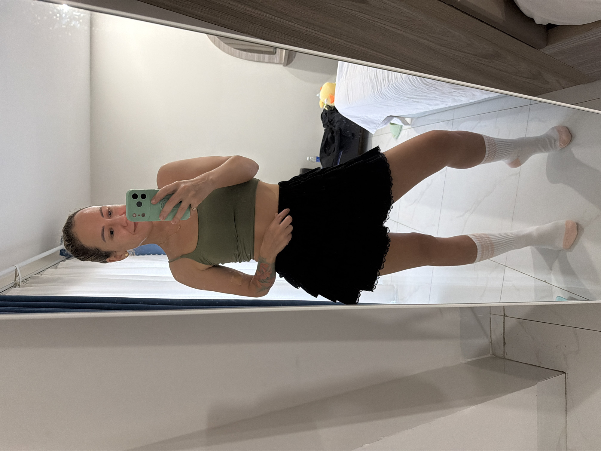 In this full-body mirror selfie, a young woman stands in what appears to be a bedroom or dressing area, with a tiled floor and light-colored walls. She is wearing a fitted olive green sports bra and a black, lacy mini-skirt. White crew socks with light pink accents cover her calves. She is holding a mint green smartphone in her right hand, which is angled to capture the reflection. Her left arm is bent, with her hand resting on her hip. She has short, dark hair pulled back and is looking towards the phone, with a slight smile on her face. A tattoo is visible on her left forearm.
In the background, a white bed with rumpled sheets is visible, along with a dark pile of clothing and a yellow plush toy. A water bottle is also on the floor. The setting appears to be indoors, possibly in a hotel room or apartment in Nha Trang, Vietnam, given the provided location context. The lighting suggests it's daytime. The overall mood is casual and relaxed, indicating the woman is likely getting ready or posing for a photo. No visible text is present in the image.