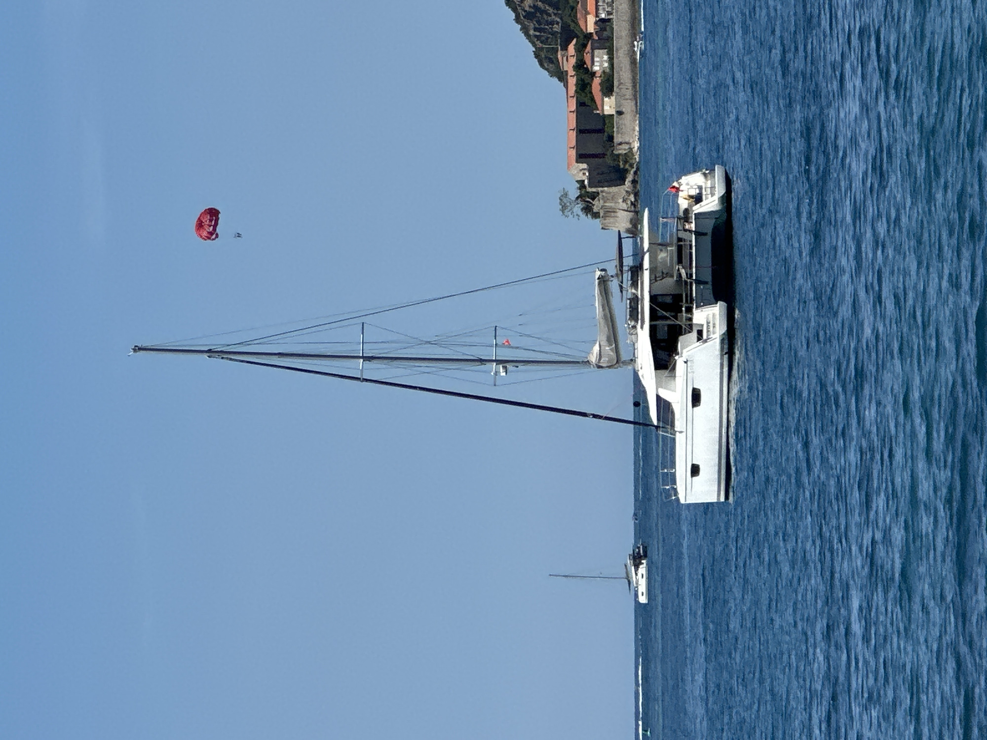The image displays a coastal scene off Budva, Montenegro, under a clear, light blue sky. A white sailing catamaran, observed from its port side and stern, is positioned in deep blue, rippled water. A red flag is visible on the catamaran's stern. In the upper right quadrant of the sky, a red parasail is aloft, with two human figures suspended beneath it. To the left, further in the distance, a white monohull sailboat is visible on the water. The right background features a rocky coastline with historical architecture characteristic of Budva's Old Town, including stone buildings with red-tiled roofs and visible fortifications. A small white powerboat, creating a wake, is present near the distant shoreline on the right. The overall scene depicts a bright, sunny day with active maritime recreation.
