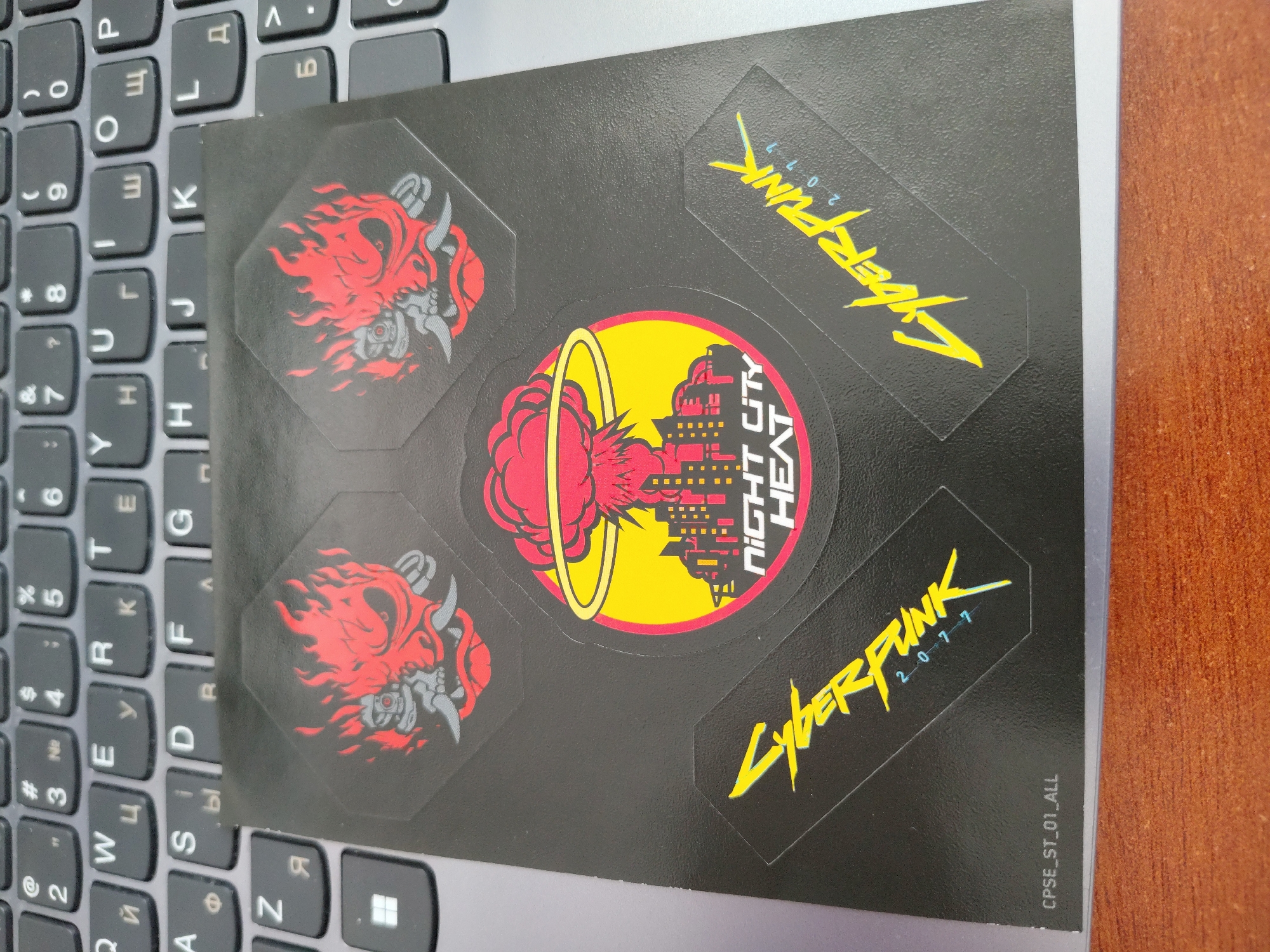 The image shows a sheet of stickers on a laptop keyboard. The stickers are black with various designs. One sticker features a red and grey demon-like mask with flames. Another is a circular sticker with a red explosion, a cityscape, and the text "NIGHT CITY HEAT" in white letters. A third sticker is yellow text that reads "CYBERPUNK 2077". There is also a small code "CPSE_ST_01_ALL" visible on the black sheet. The laptop keyboard is dark grey and appears to be a standard QWERTY layout with some Cyrillic characters visible. The setting is likely indoors, given the presence of a laptop and the lighting. No people are visible in the image.