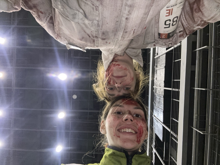 A close-up, inverted-perspective photograph captures two smiling individuals covered in simulated blood against an industrial-like background in Huangdao District, China.
In the foreground, an adult female is visible from the upper torso upwards. She wears a white, long-sleeved garment, likely a jumpsuit, heavily stained with red liquid consistent with simulated blood. A rectangular white patch is affixed to her left shoulder, displaying the numbers "3", "85", and the letters "IE", alongside an unreadable barcode. Her face, framed by light-colored hair, is also smeared with simulated blood. She exhibits a slight smile and directs her gaze toward the camera.
Positioned behind the first individual, an adult female's head and upper shoulders are visible. She wears a green-colored upper garment. Her face is extensively covered in simulated blood, and she displays a wide smile while looking directly at the camera. Her hair is dark.
Both individuals are actively posing for the photograph, making direct eye contact with the camera, which suggests a non-distressed demeanor despite the simulated injuries.
The background is dark. To the left, a structure composed of dark horizontal and vertical bars or grates is partially visible. To the right, multiple bright, circular light sources are discernible against a dark, possibly ceiling-like surface, casting glare. The overall environment suggests an indoor setting, potentially an industrial facility, soundstage, or theatrical production area. The scene is located in Huangdao District, China.