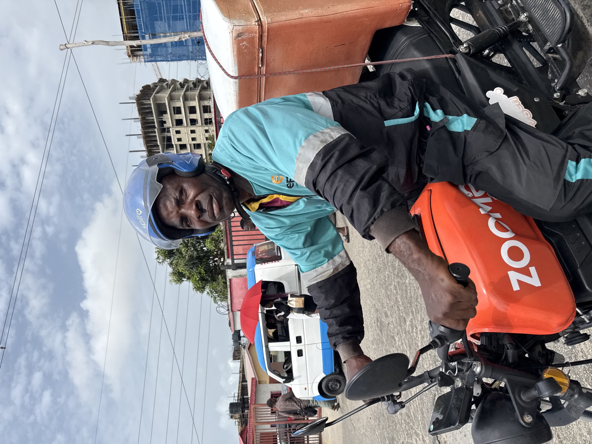 A medium shot captures a motorcycle rider in Itirin, Nigeria. The rider, wearing a light blue helmet, a turquoise and black jacket, and displaying visible signs of fatigue or weariness, is seen seated on his motorcycle. The motorcycle features an orange ZOO delivery box mounted to its rear. In the background, urban elements are visible, including a minibus with a red umbrella near its doorway, and a building under construction framed against a partly cloudy sky. The photo seems to have been taken at ground level during daylight, portraying a scene of everyday activity in the city.