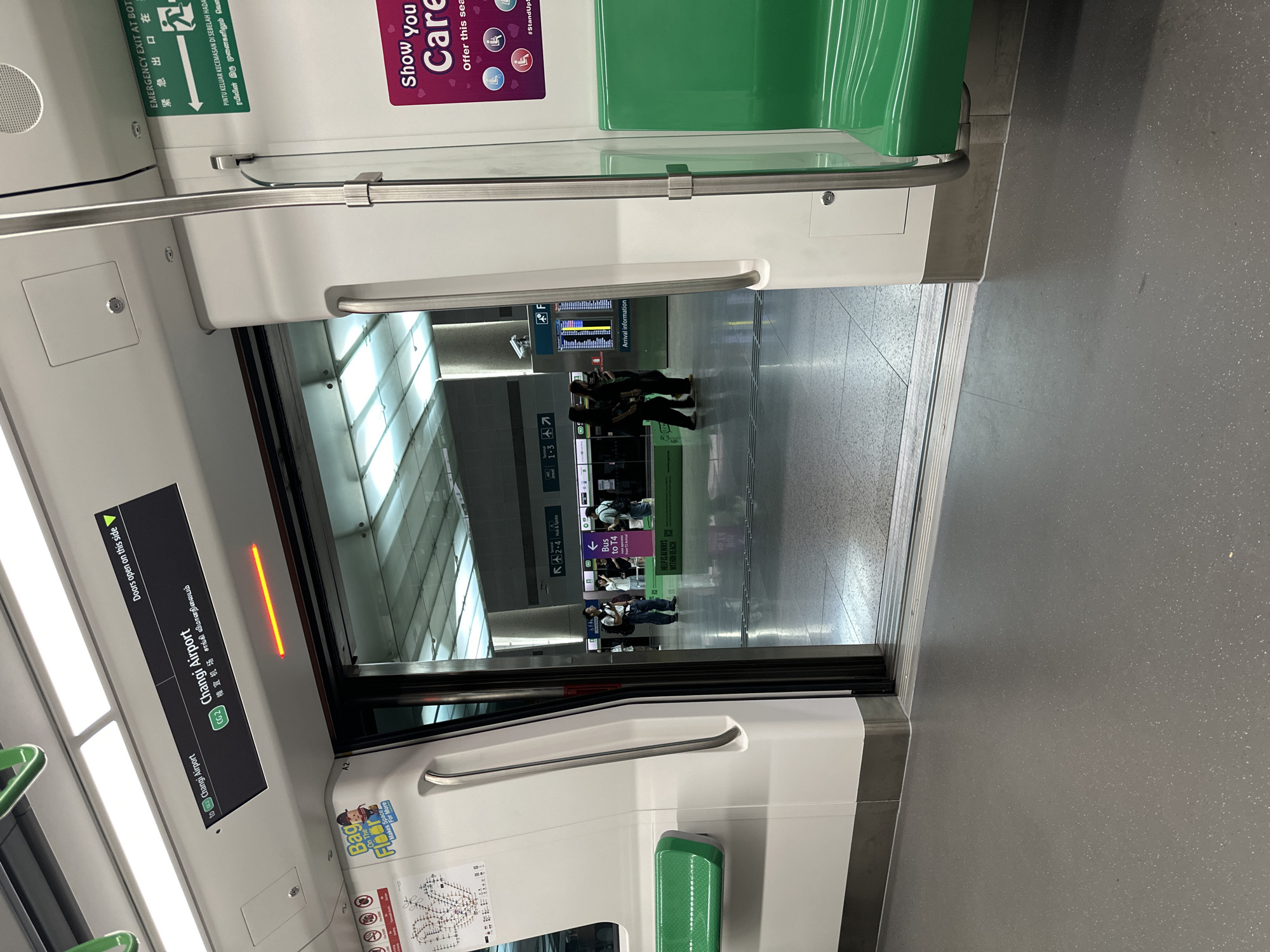 The image is taken inside a Singapore MRT train carriage, looking out through the open doors onto a station platform. The train carriage interior is white with green seats and metal grab bars. A digital sign above the doors indicates the train is heading to "Changi Airport" (CG2) and displays "Doors open on this side" in English, Chinese and Tamil. On the platform, several people are visible, some walking and others standing. One person is looking at an arrival information screen. The station has a modern, clean aesthetic with tiled floors and a high ceiling with ample lighting. A sign on the wall of the carriage advertises "Show you Care" with icons representing elderly, pregnant, and disabled individuals, suggesting the priority seating policy.