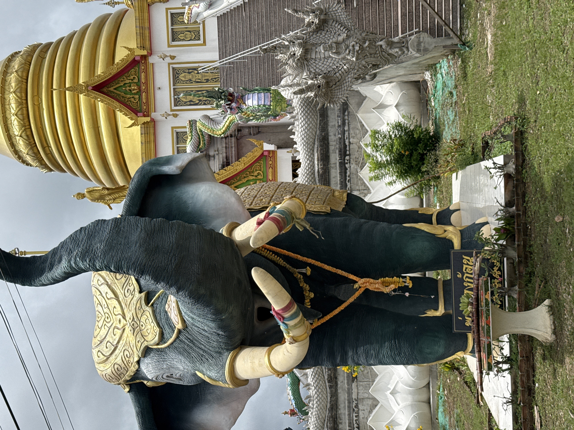 The image depicts a large, dark green elephant statue in the foreground, adorned with a golden decorative headpiece and ivory tusks. The statue is decorated with colorful bands around its tusks and a floral garland around its neck. Behind the elephant, a white building with a golden tiered roof, characteristic of a Thai temple, is visible. A grand staircase with stone serpent balustrades leads up to the temple entrance. The scene is set outdoors, with a cloudy sky overhead. A sign with Thai script is placed in front of the elephant statue, and on a small table in front of the sign, sugarcane stalks and bananas are arranged as an offering. The location is identified as Wat Khun Inthapramun, Ang Thong Province, Thailand.