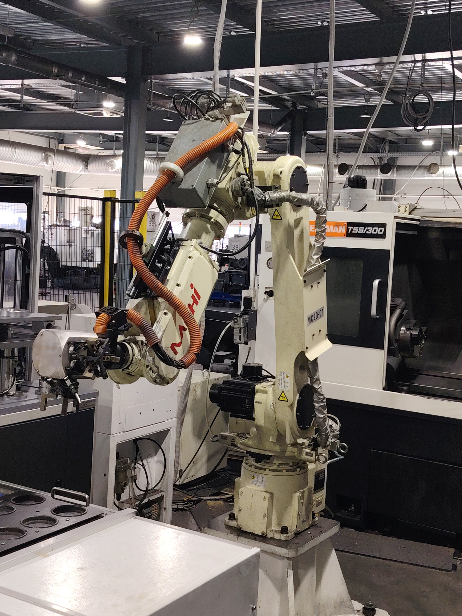 The image shows an industrial setting with a FANUC robot, identified by the "FANUC" lettering on one of its arms. The robot appears to be integrated with a machine tool, labeled "LEHMANN T55/300," likely a CNC lathe or milling machine. The setting suggests a factory or manufacturing plant, with overhead lighting and support structures visible. A metal fence is present in the background, possibly separating different work areas. The robot's arms are covered in protective conduits. There are no people visible in the frame. There is no indication or evidence of this being in the city of Noviy, Russia.