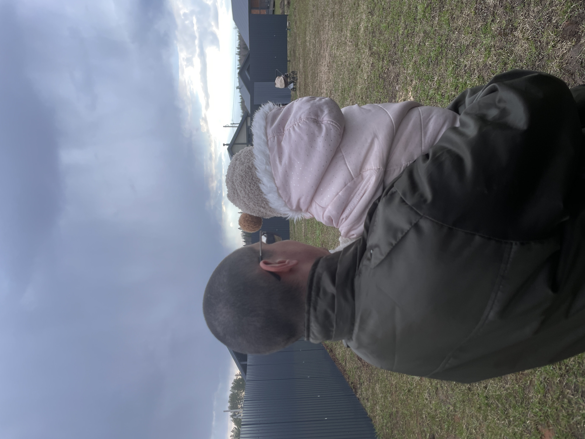 The image captures an outdoor scene in Подшивалово, Russia, featuring a man and a small child. The man, viewed from behind, has a closely-shaved head and is wearing a dark green jacket. A baby or toddler is positioned on his back or shoulders, dressed in a light pink puffy winter coat with a furry white hood and a light brown, ear-flapped hat adorned with small pompoms. Both appear to be looking forward, seemingly observing the landscape in front of them.
The setting is an open, grassy area with sparse and patchy green and brown grass, suggesting either early spring or late autumn. In the mid-ground, several modern, dark grey or black buildings with sloped roofs are visible, resembling houses or cabins with contemporary architectural styles. One building features a corrugated metal or panel exterior. A stroller or wagon-like object is partially visible between two of these structures. A line of trees can be seen in the far distance, close to the horizon.
The sky is predominantly overcast with heavy grey clouds, though a brighter section on the horizon suggests the sun breaking through or rising/setting. The diffused light and warm clothing worn by both subjects indicate a cool or chilly day. The overall activity appears to be a calm, observational moment outdoors. No visible text is present in the media file.