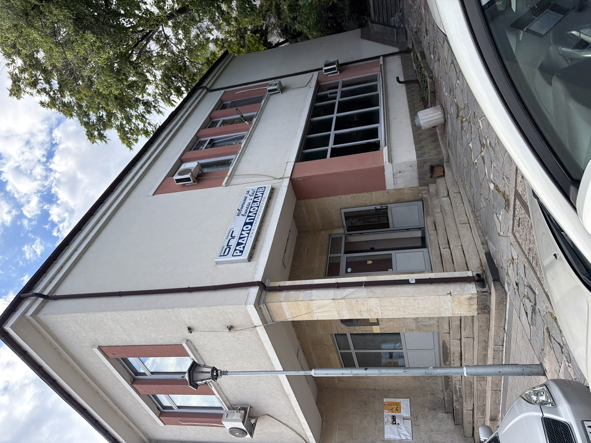 The image depicts the exterior of the Bulgarian National Radio (BNR) Plovdiv building, located in the city of Plovdiv, Bulgaria. The building is a two-story structure with a light-colored facade accented with pinkish-red rectangular sections around some windows and at the corners. It features several windows, some with vertical decorative elements, and air conditioning units are visible on the exterior walls. A prominent sign with the letters "BNR" and the text "РАДИО ПЛОВДИВ" (Radio Plovdiv) is mounted on the building. Below this, a website address "radioplovdiv.bg" is also visible.
To the left of the entrance, there is a vintage-style street lamp. A series of stone steps leads up to two entrances on the ground floor. On the left side of the building, there are bulletin boards with posters. The foreground shows the hoods and windshields of two parked cars, one silver and one white. The ground in front of the building is paved with irregular stone slabs, some of which appear cracked. Vegetation, including a large tree with dense green foliage, is visible in the background on the right side of the image. The sky above is partly cloudy with patches of blue. The overall scene suggests an urban environment with a focus on the radio station's building.