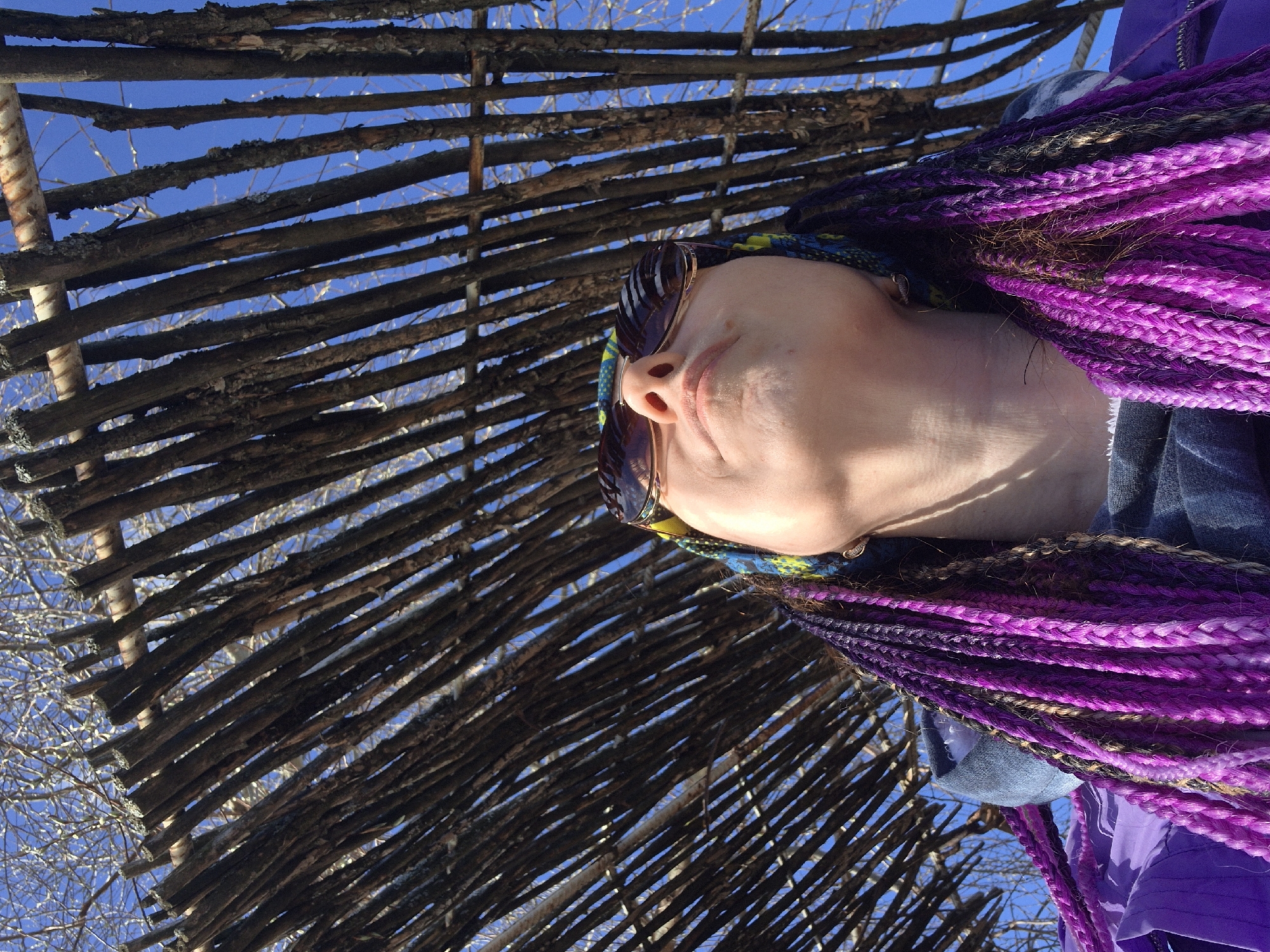 A low-angle shot captures a person with purple-braided hair wearing sunglasses, positioned in front of a rustic, stick-built fence. The background reveals a clear blue sky, sparsely interspersed with bare tree branches. The location appears to be an outdoor setting in the city of Chaikovsky, Russia, based on geographical data.